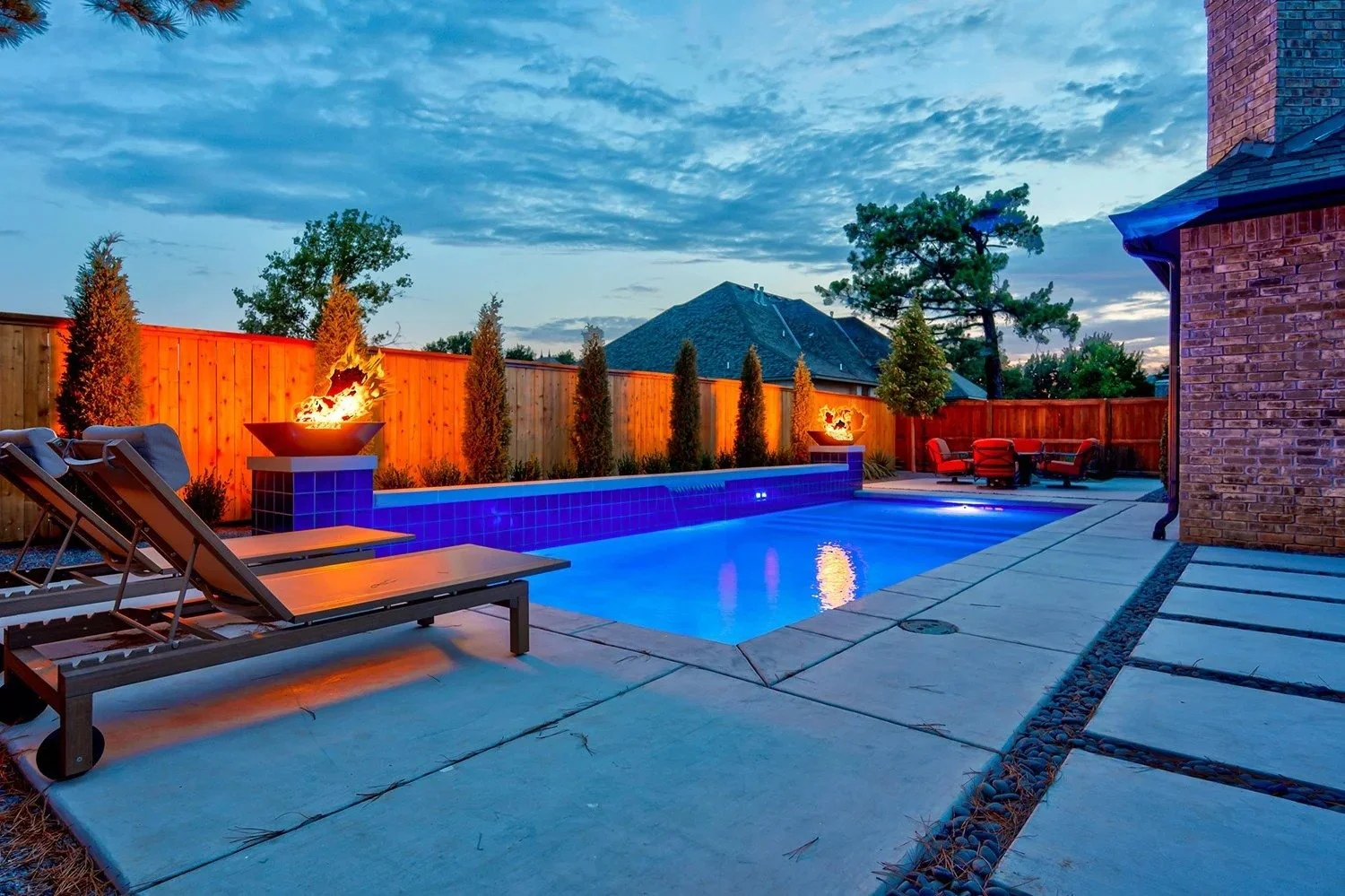 Backyard with illuminated swimming pool, outdoor seating, and decorative fire features during twilight.