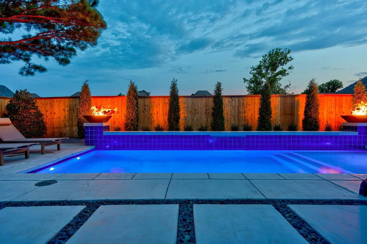 A backyard swimming pool illuminated with blue lights during dusk, surrounded by a concrete patio with two lounge chairs on the left, a wooden privacy fence, and tall bushes and trees along the fence. Two fire bowls with flames are placed on pedestal