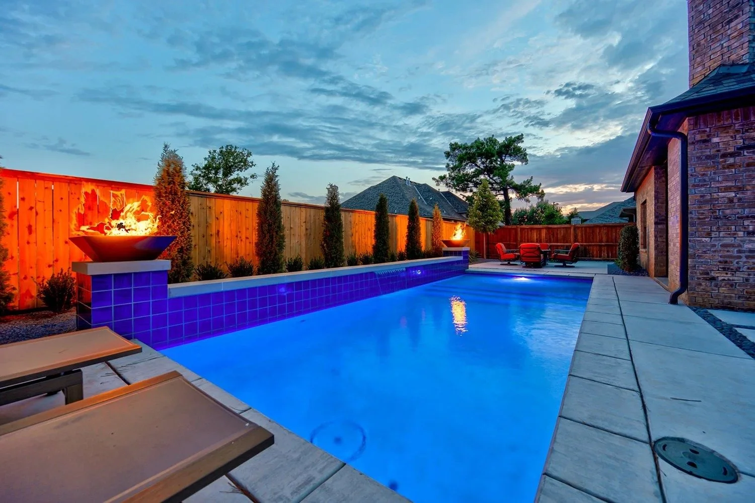 Backyard with illuminated swimming pool, wooden fence, outdoor seating with red chairs, fire bowls, trees, and cloudy sky at dusk.