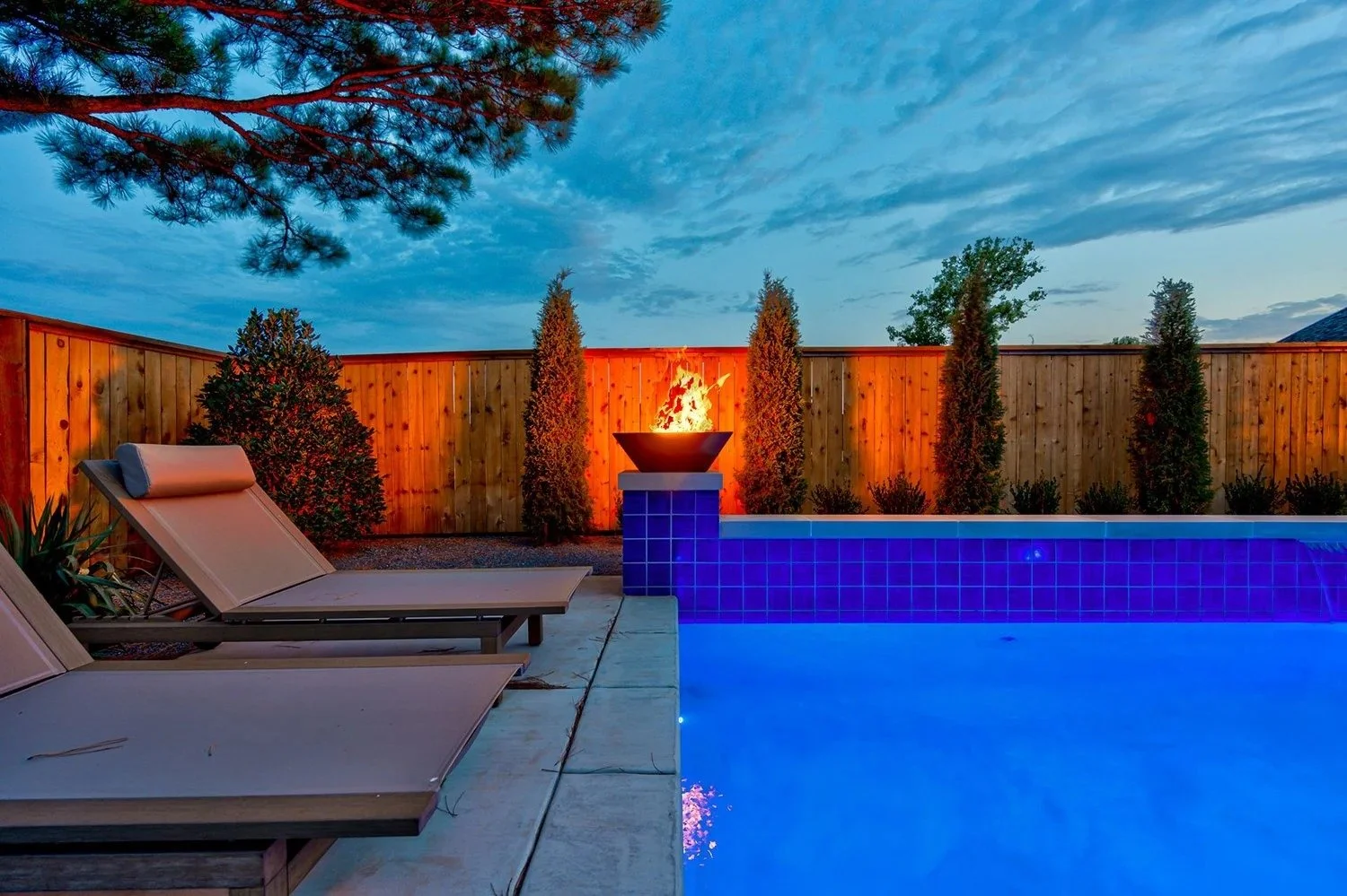 A backyard pool area at dusk with two lounge chairs, a fire feature, tall trees, and a wooden privacy fence under a partly cloudy sky.