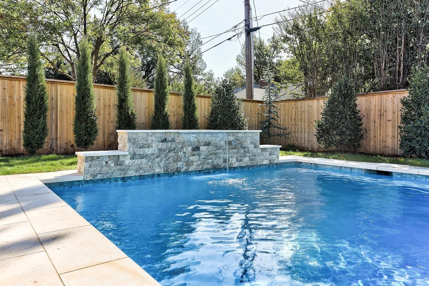 Backyard swimming pool with a stone waterfall feature, surrounded by a wooden fence and tall green trees.