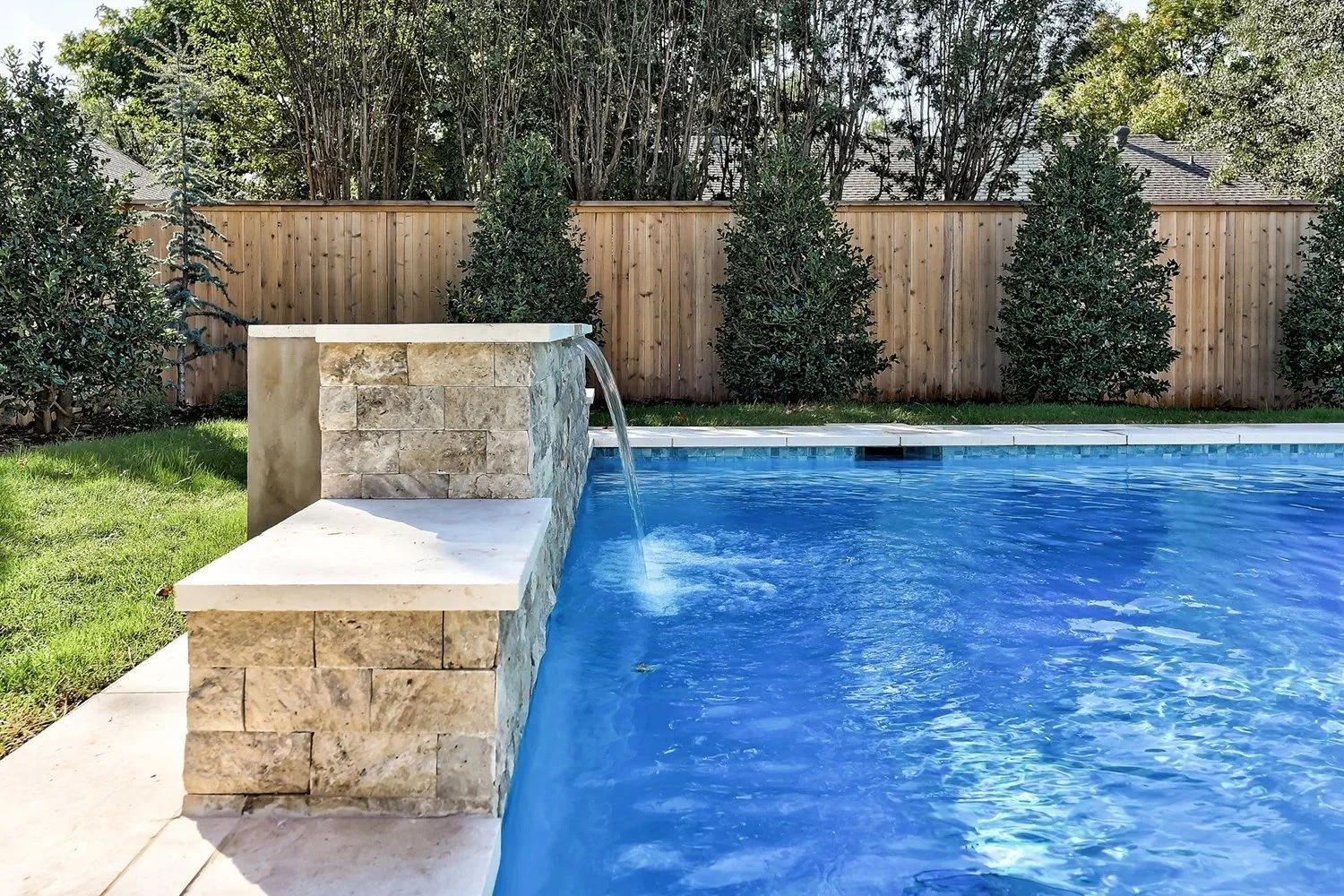 Backyard swimming pool with a stone water feature and a wooden fence, surrounded by bushes and trees.