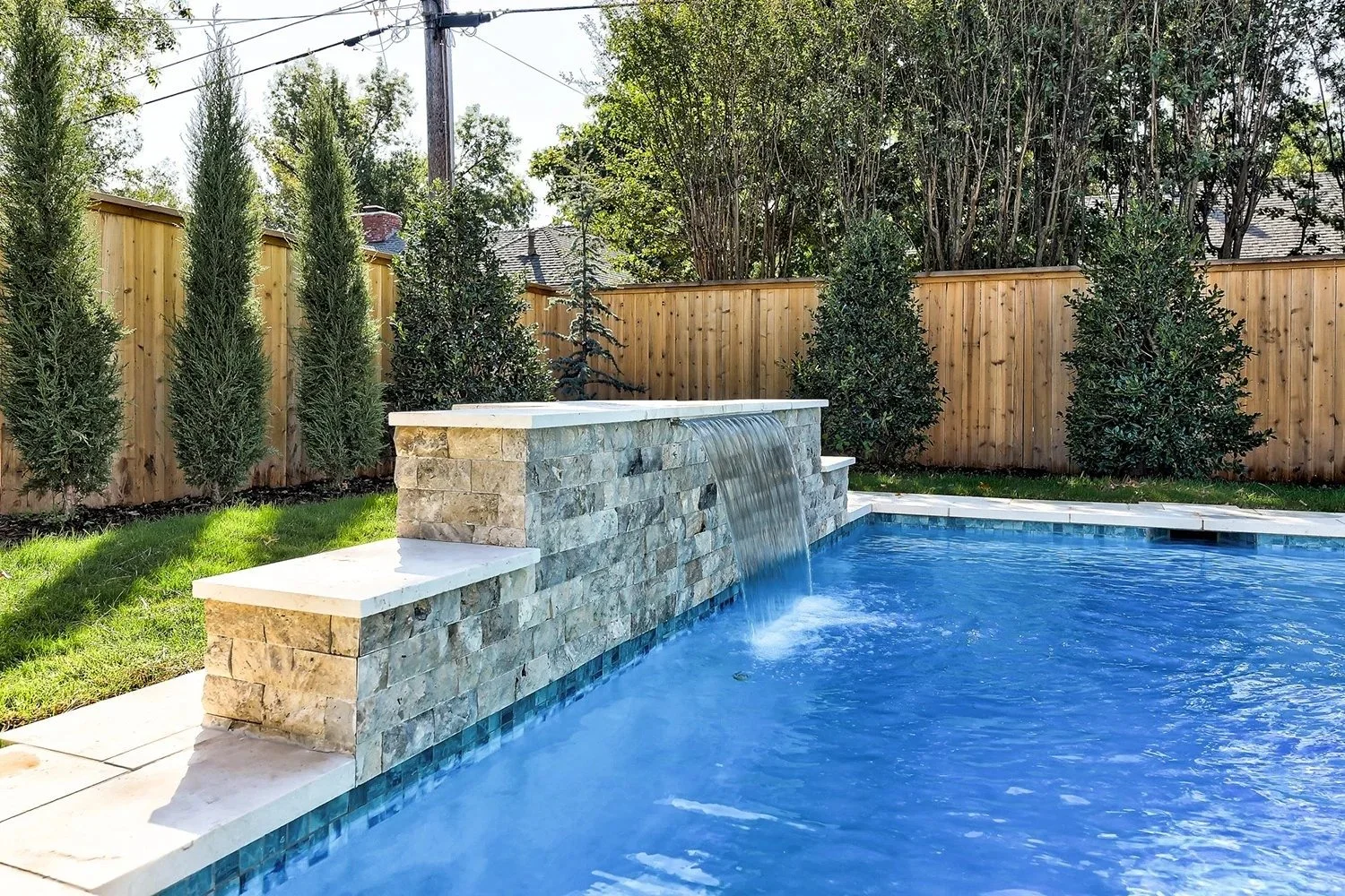 Clean backyard swimming pool with waterfall feature, surrounded by a wooden fence and trees.