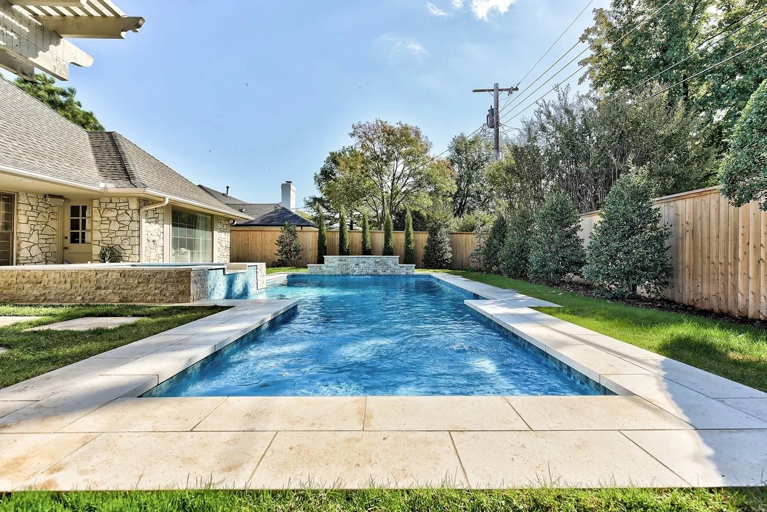 A backyard with a rectangular swimming pool, a waterfall feature at the far end, and a stone patio surround. There is a house with stone walls and a large window on the left, a wooden fence on the right, and trees providing greenery along the fence l