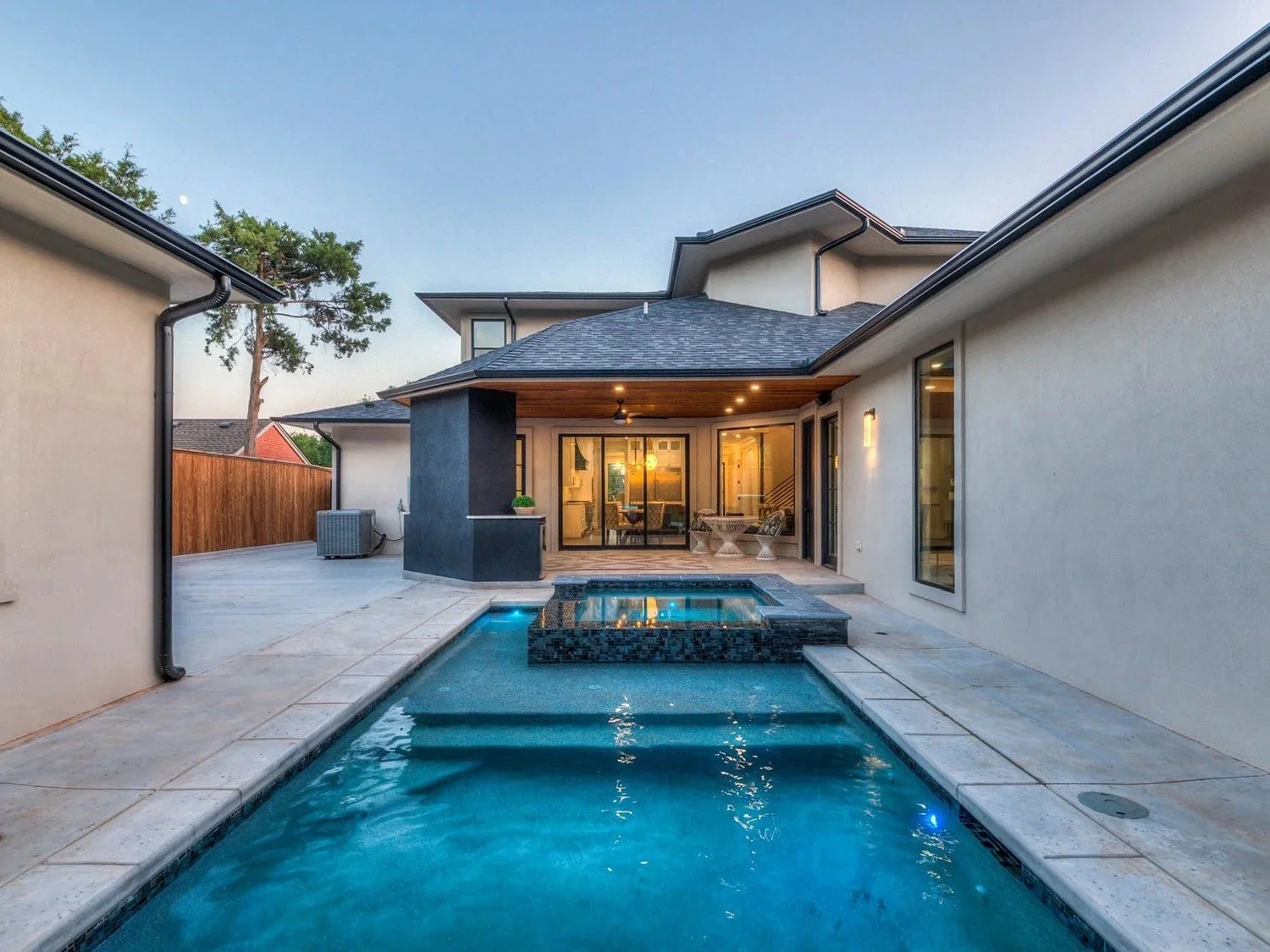 Backyard with swimming pool and hot tub, outdoor seating area, house with large windows and lights on, wooden fencing, trees, clear sky at dusk.