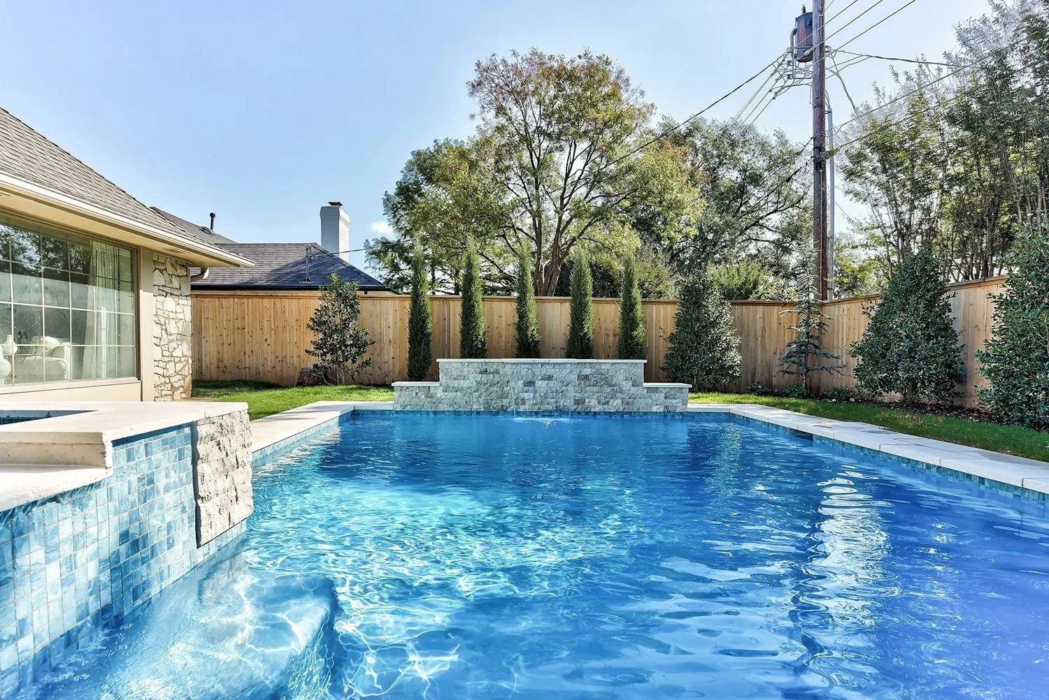 Backyard with a rectangular swimming pool, stone waterfall feature, and wooden fence, surrounded by trees and greenery.