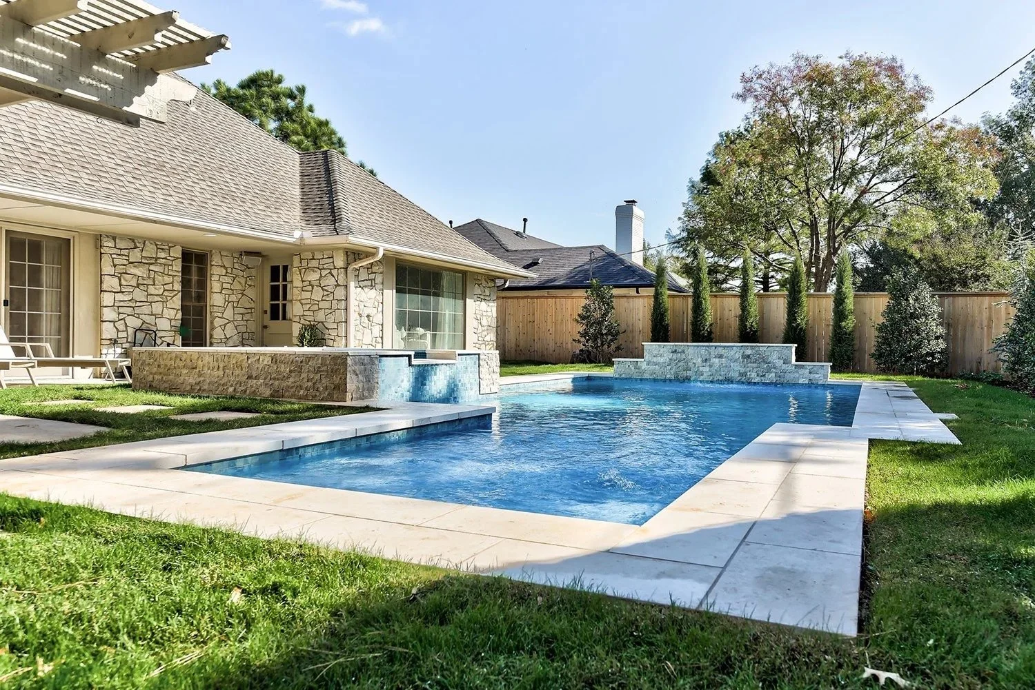 Backyard with swimming pool and stone house wall, sitting area with lounge chairs, wooden fence, tall trees, and lush grass.