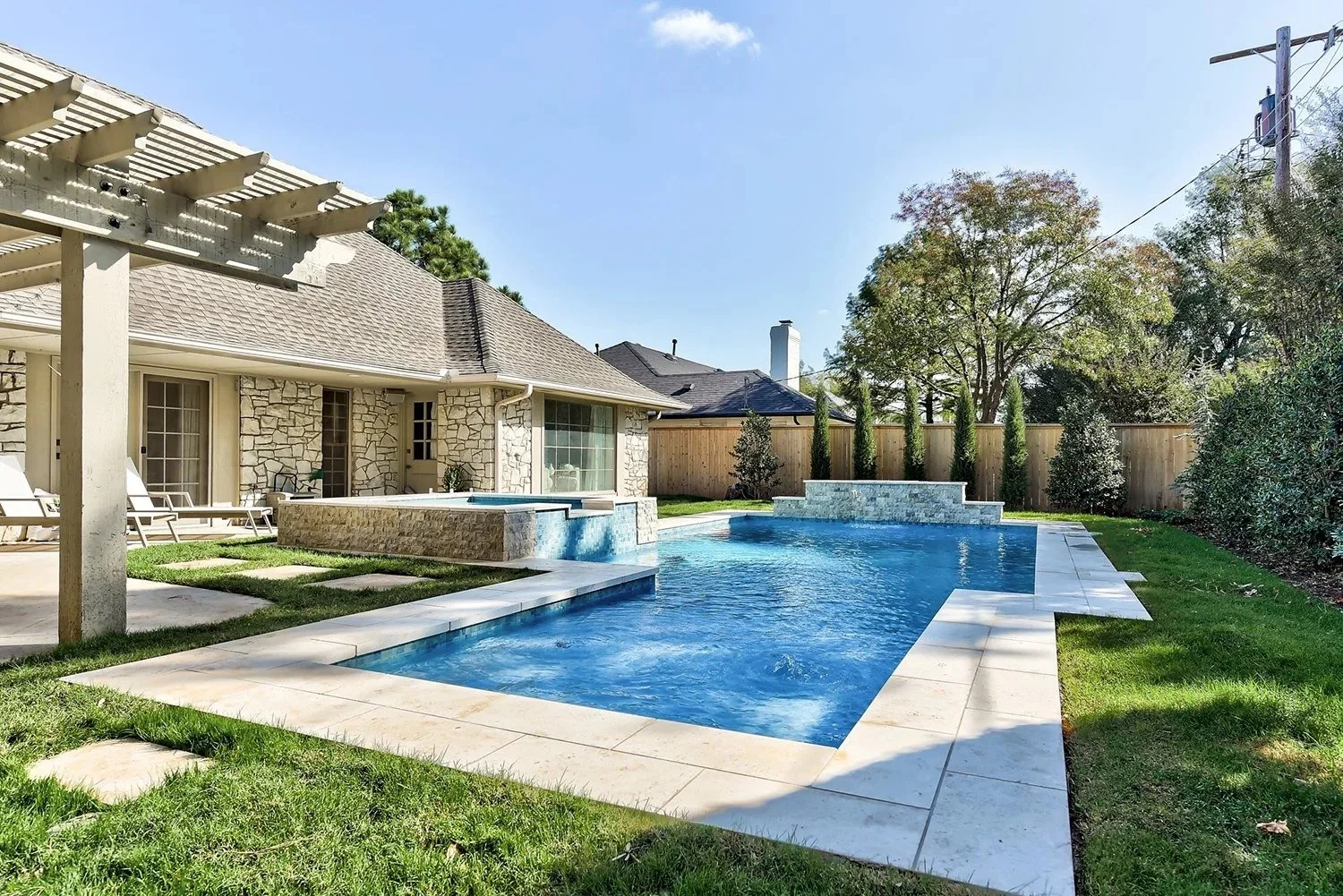 Backyard with a swimming pool, stone patio, lounge chairs, house with stone exterior, surrounding trees and shrubs, and a wooden fence.