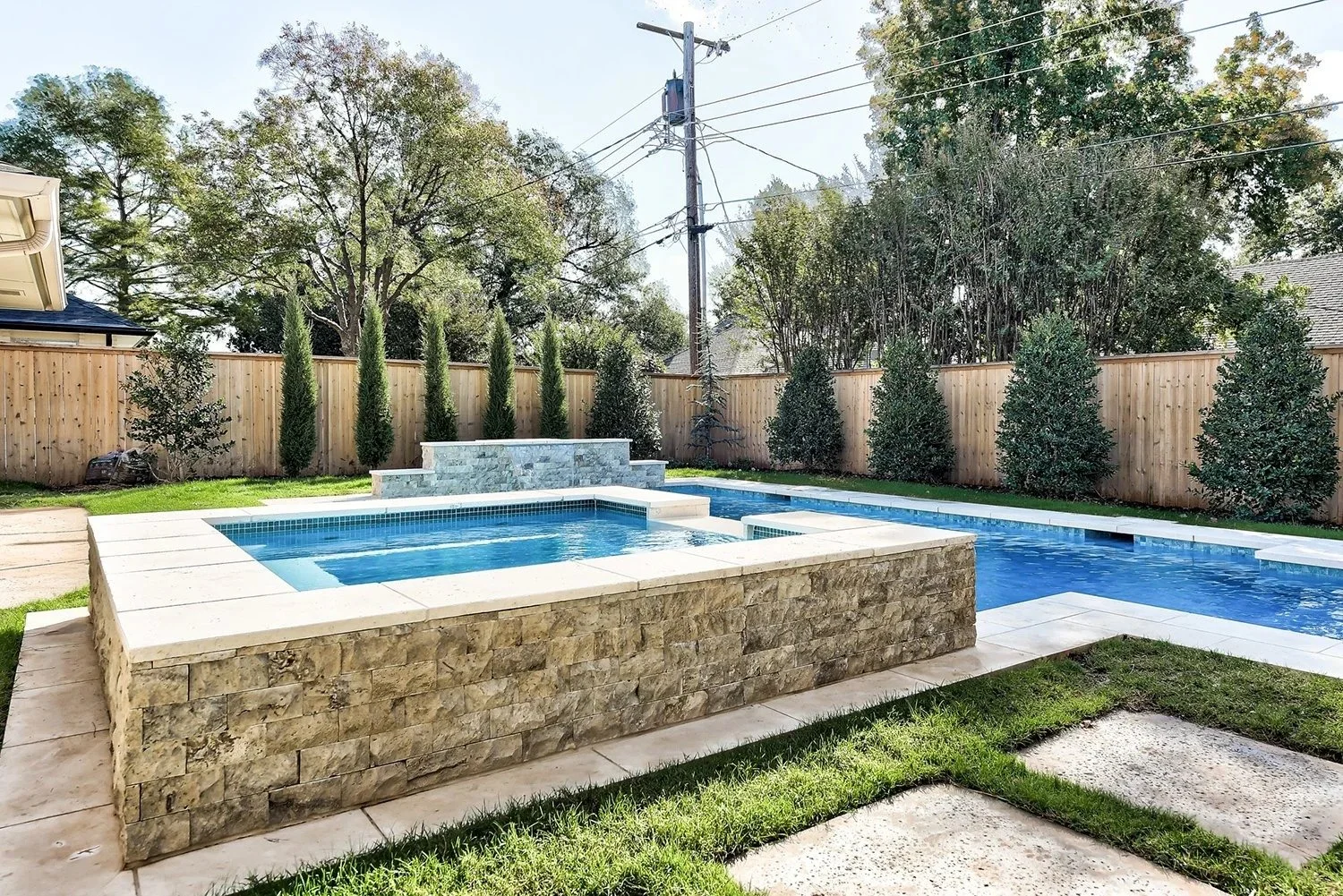 Backyard with a small pool and a stone spa, enclosed by a wooden fence and surrounded by trees and shrubs.