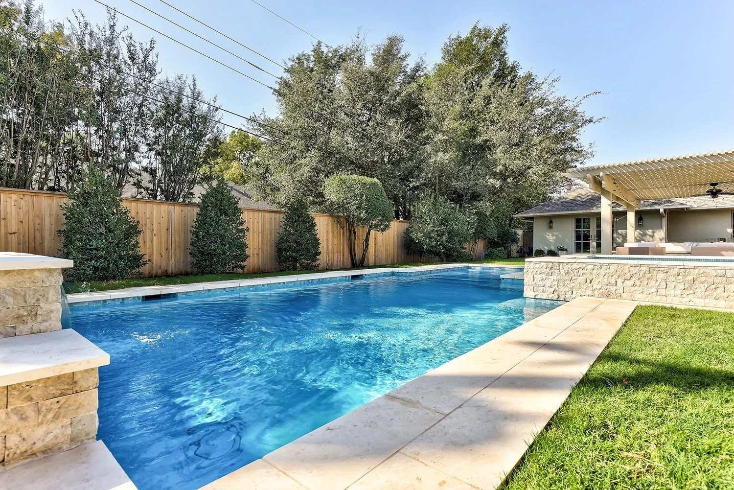 A backyard with a rectangular swimming pool, a wooden fence, green trees, a grassy lawn, and a covered patio area with outdoor furniture.