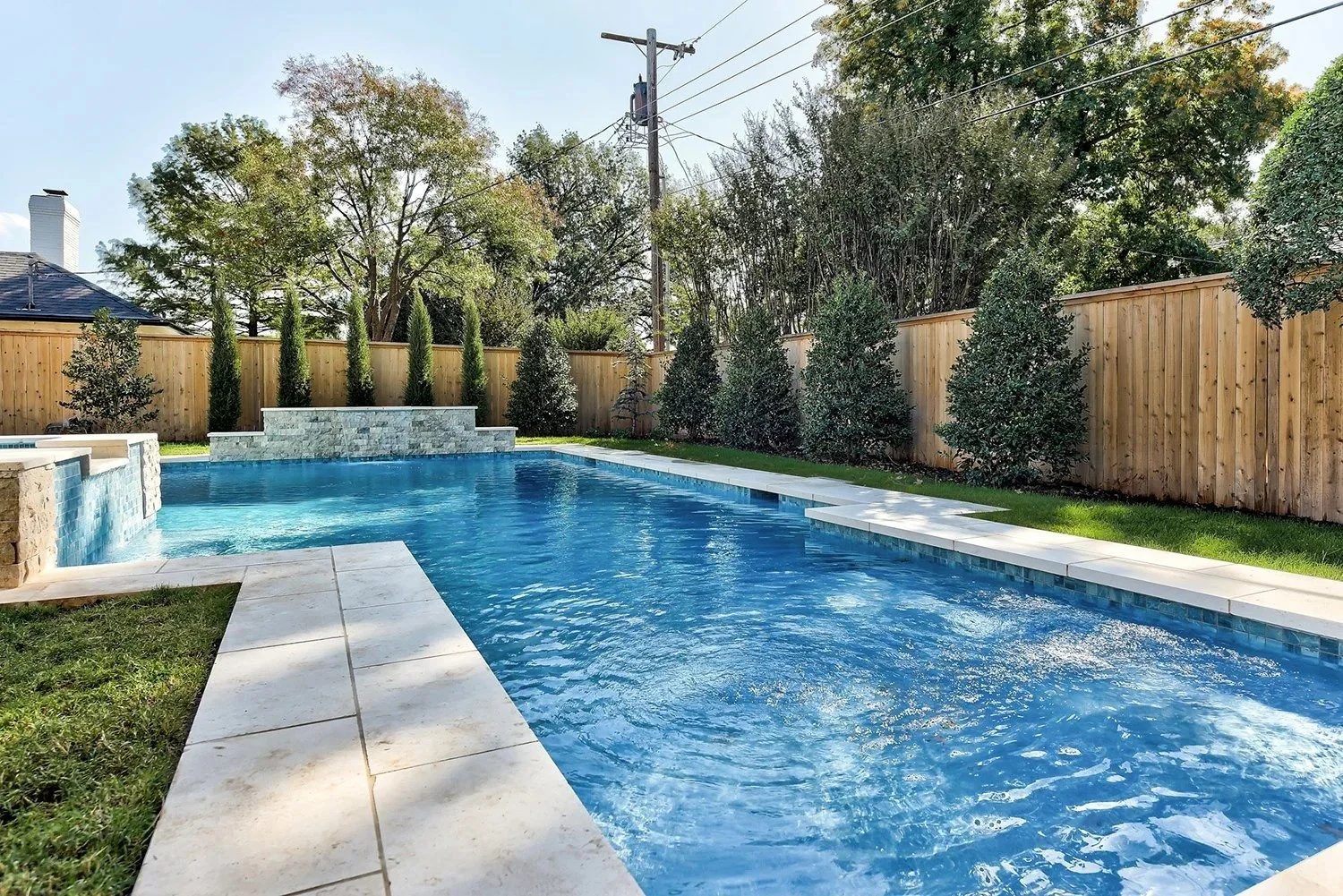 Backyard with a swimming pool, stone edging, a wooden privacy fence, and trees in the background during daytime.