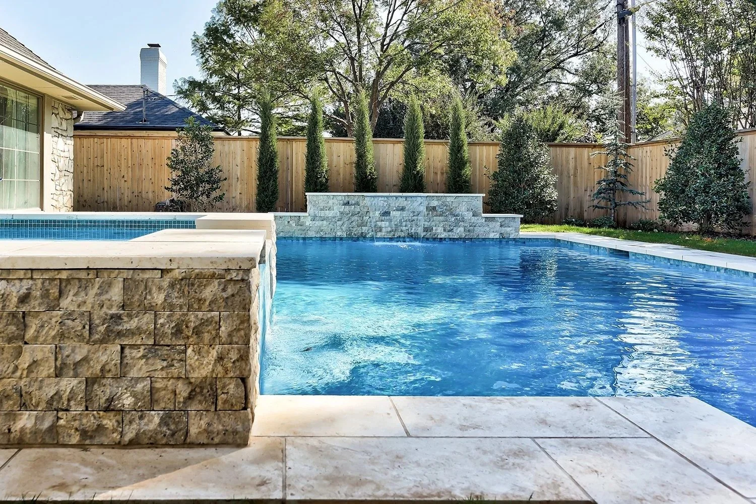 Backyard swimming pool with stone edge, surrounded by a wooden privacy fence, tall evergreen trees, and a house with stone siding.
