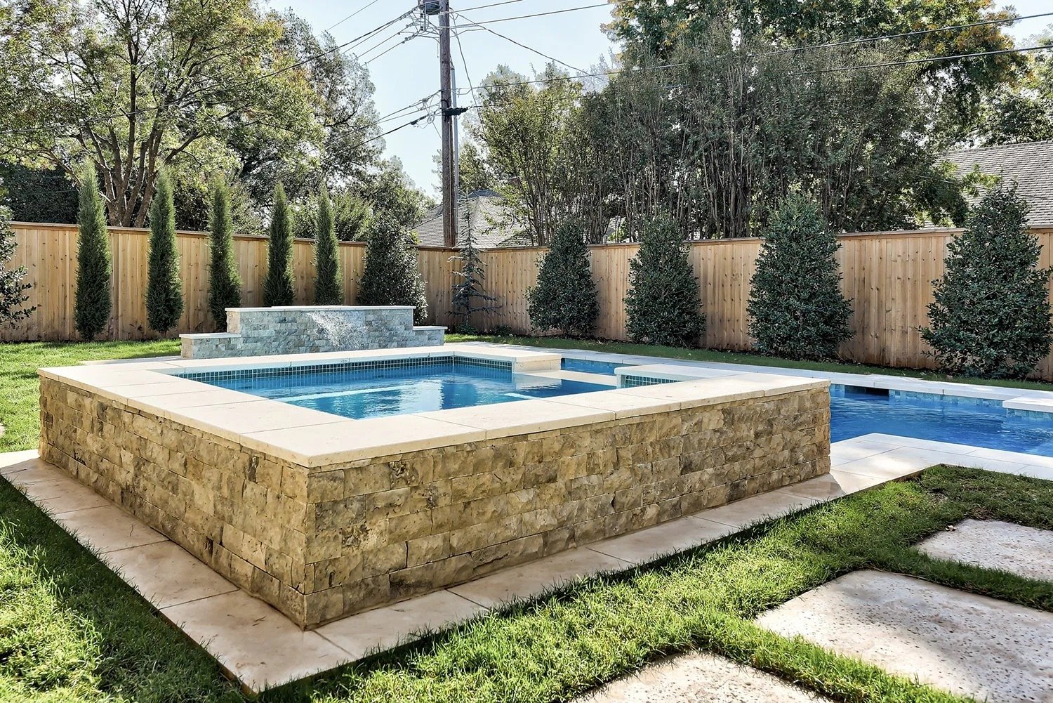Backyard with a pool and hot tub enclosed by a wooden fence, with trees and greenery.
