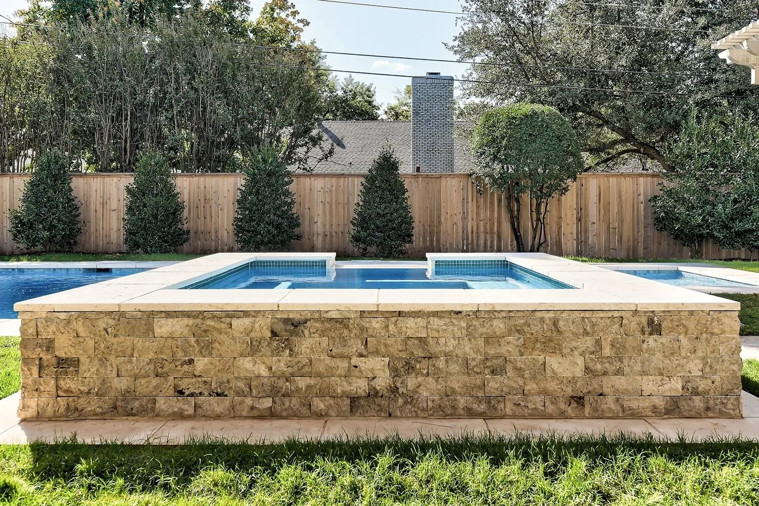 Backyard with a swimming pool and hot tub, a wooden fence, trees, and shrubs for privacy.