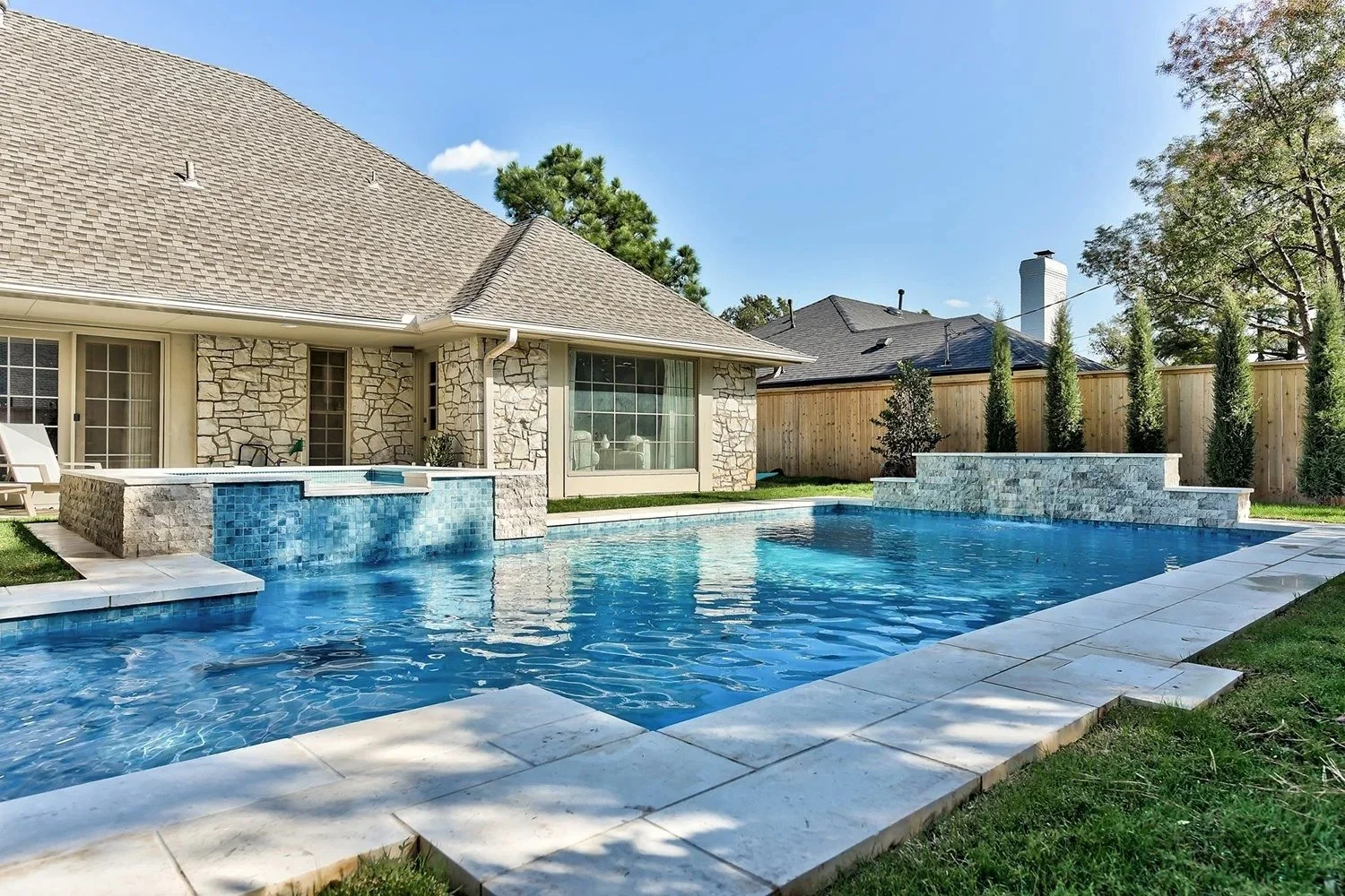 Backyard with a swimming pool, stone house, small waterfall feature, and a wooden privacy fence with trees.