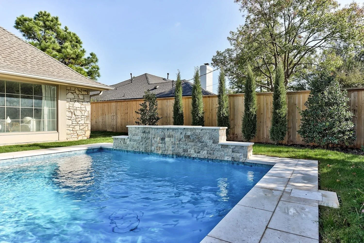 Residential backyard with a rectangular swimming pool, stone waterfall feature, green grass, tall trees, and a wooden fence, with neighboring rooftops visible.