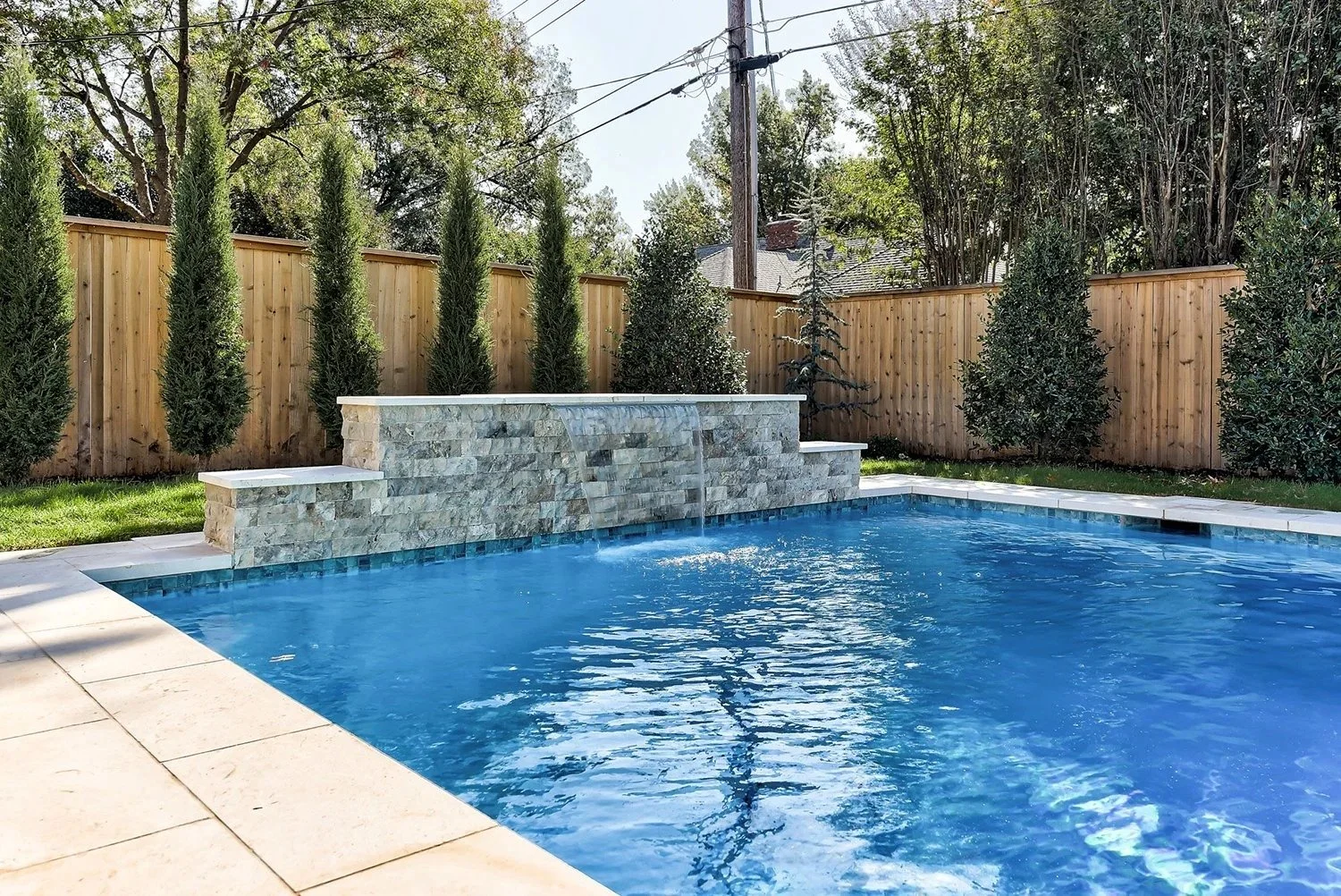 A backyard swimming pool with a stone waterfall feature, surrounded by a wooden privacy fence and tall trees.