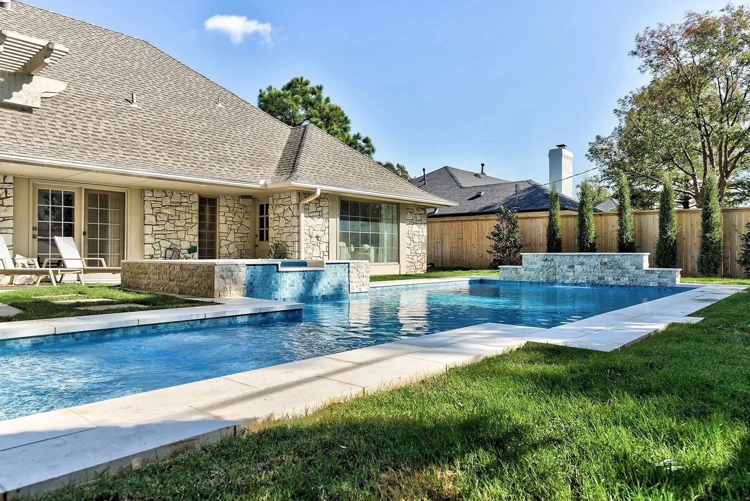 Backyard with swimming pool, stone patio, lawn, and house with stone exterior and large windows.