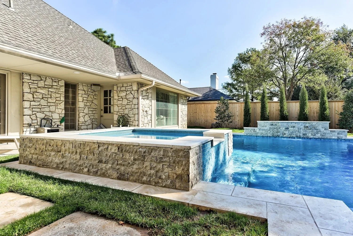 Backyard with a large swimming pool, hot tub, and stone house with a gabled roof, green lawn, trees, and wooden fence.