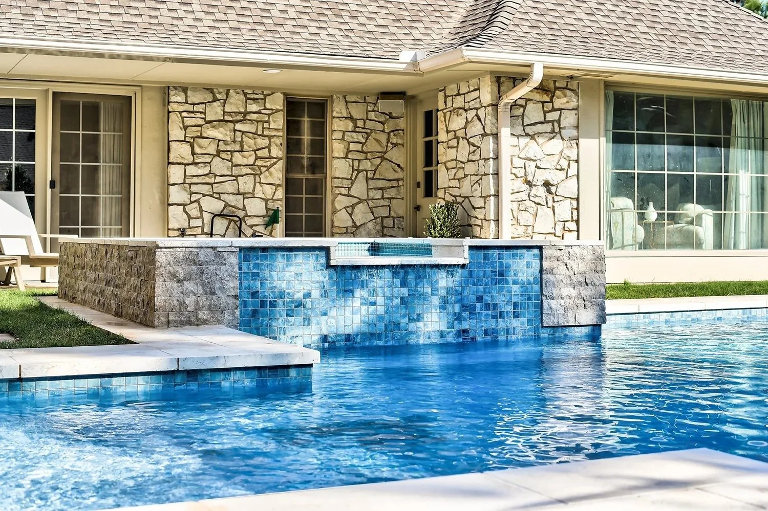 Backyard pool with blue mosaic tiles and stone border, house with large windows, stone exterior wall, and patio furniture in the background.