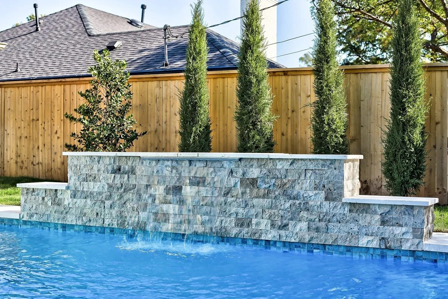 Backyard swimming pool with a stone water feature and tall cypress trees in front of a wooden privacy fence.