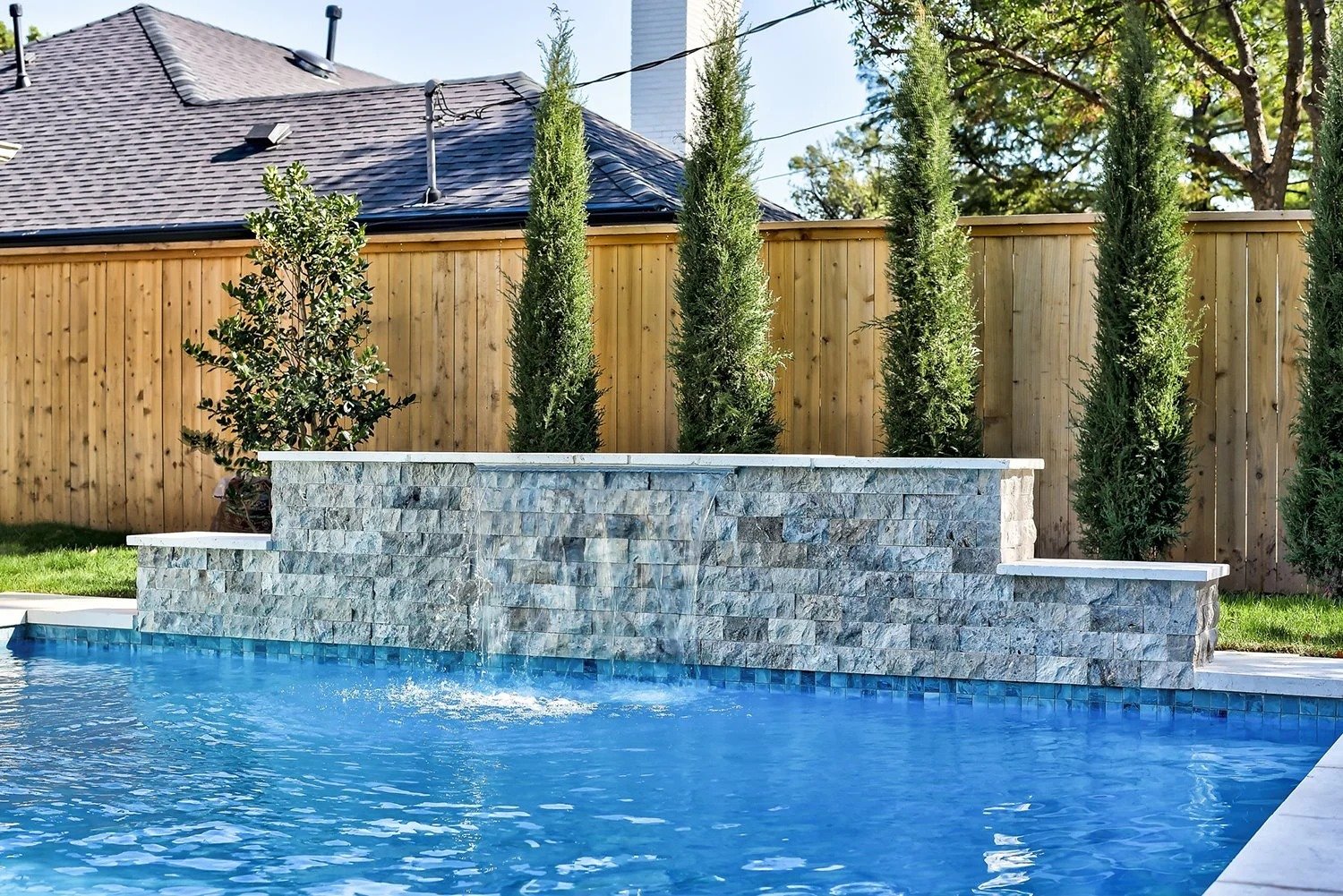 A backyard swimming pool with a stone waterfall feature, tall evergreen trees, and a wooden privacy fence. A house with a shingled roof is in the background.