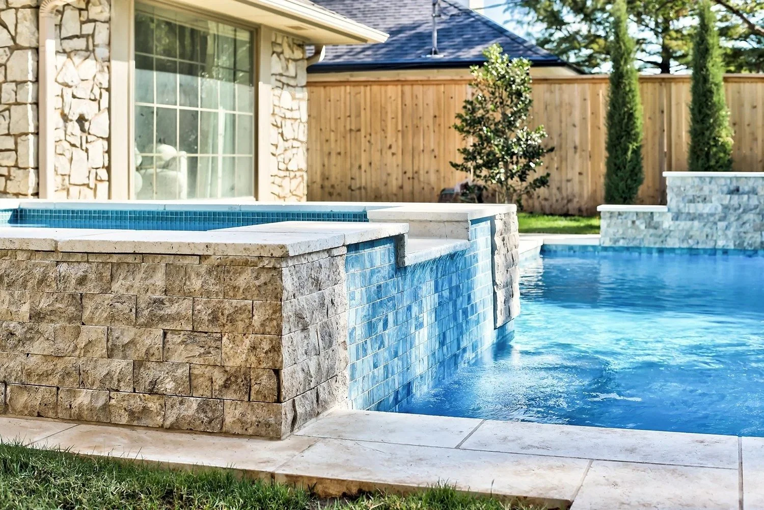 A backyard with a swimming pool and spa with blue tile accents, stone walls, a wooden privacy fence, and a glass sliding door to the house. Green grass and a few small trees are also visible.