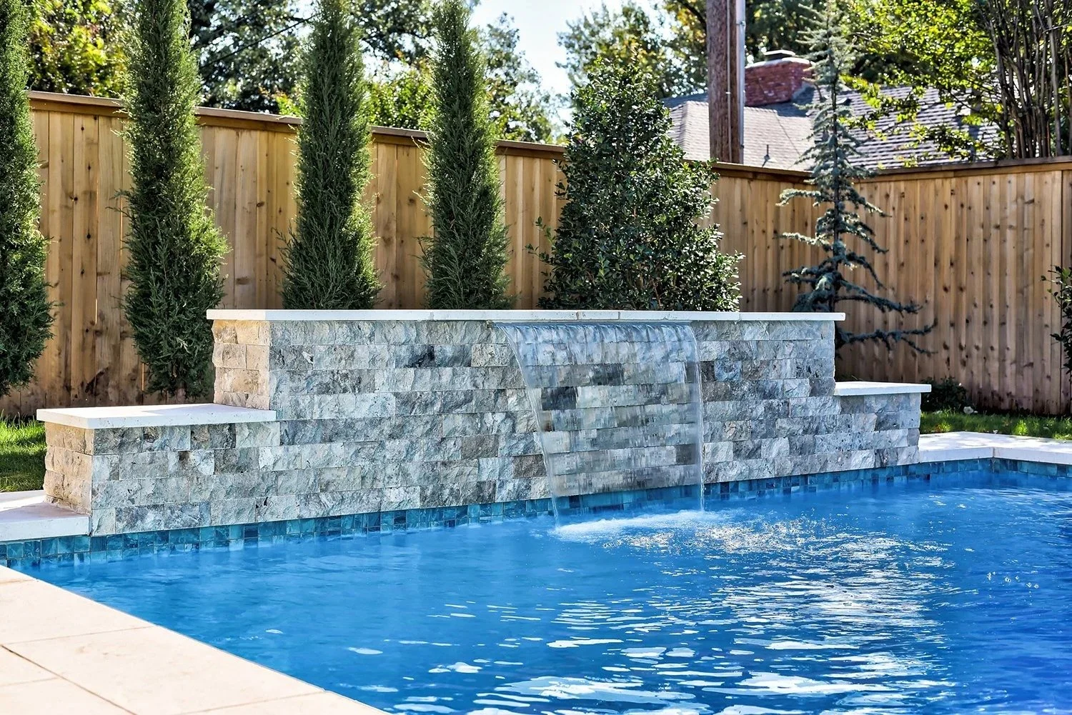 Backyard pool with a stone waterfall feature, surrounded by a wooden fence and tall evergreen trees.