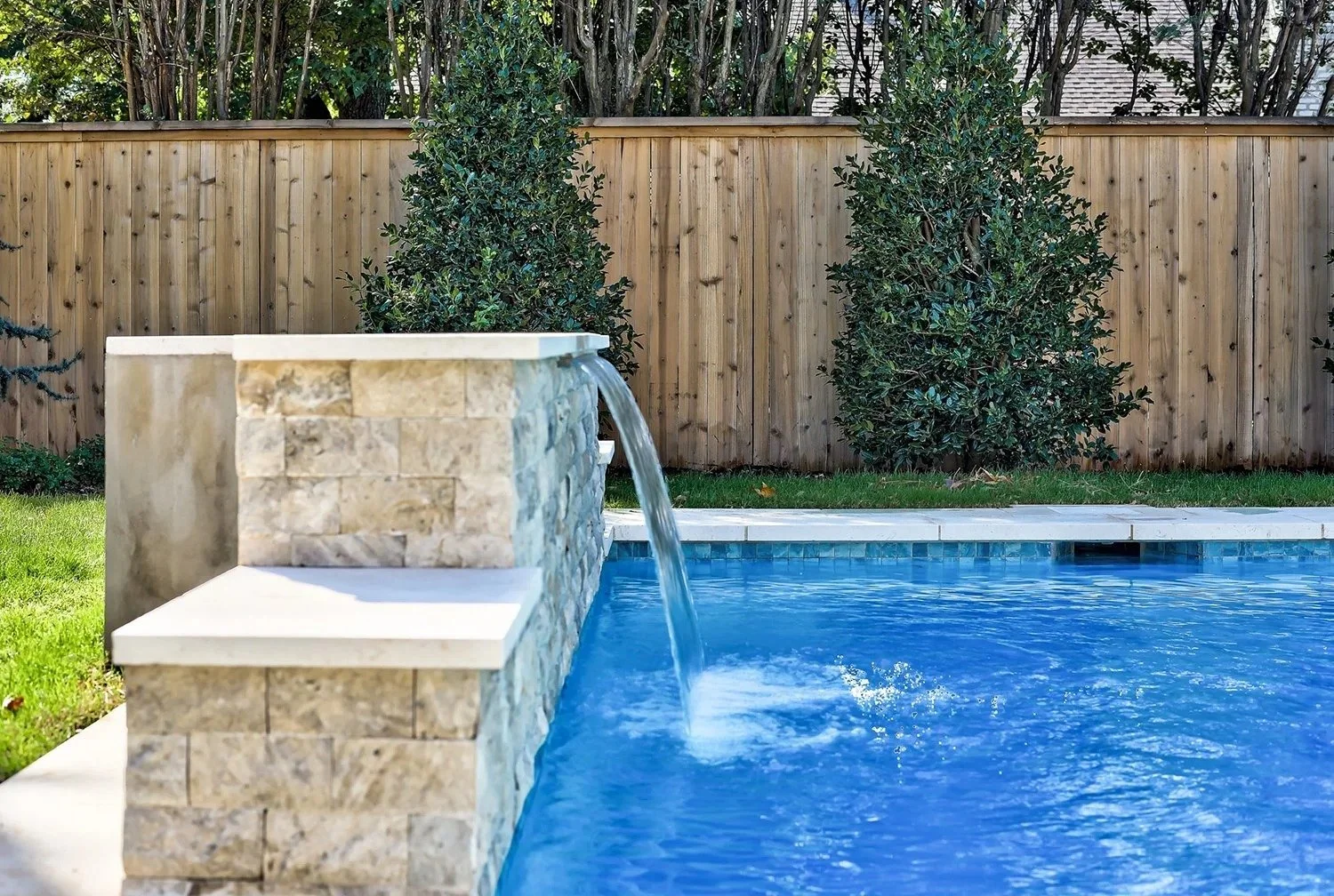 A backyard swimming pool with a stone water feature, a wooden fence, and two bushes in the background.
