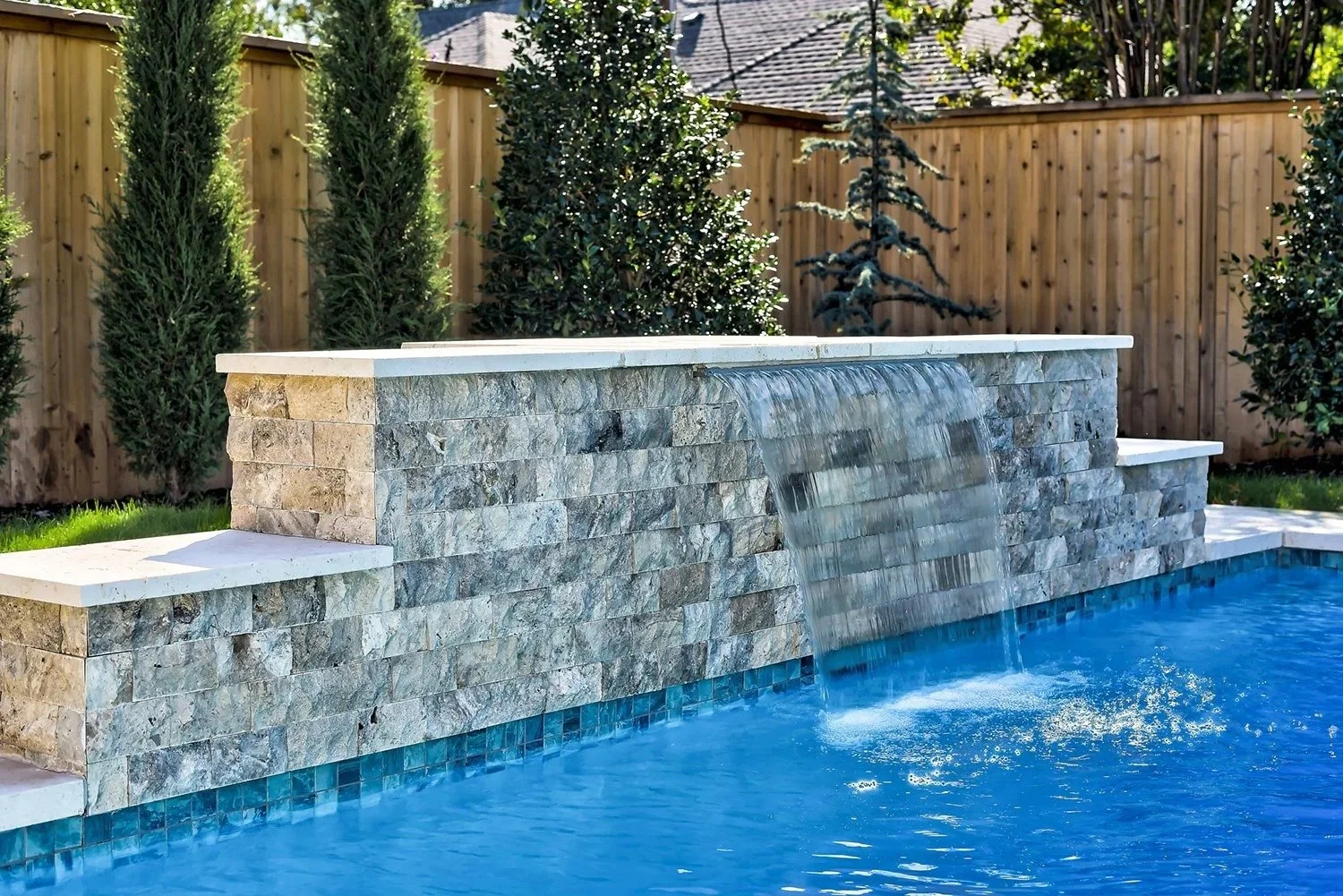 A backyard pool with a stone water fountain feature with water cascading into the pool, surrounded by green trees and a wooden fence.