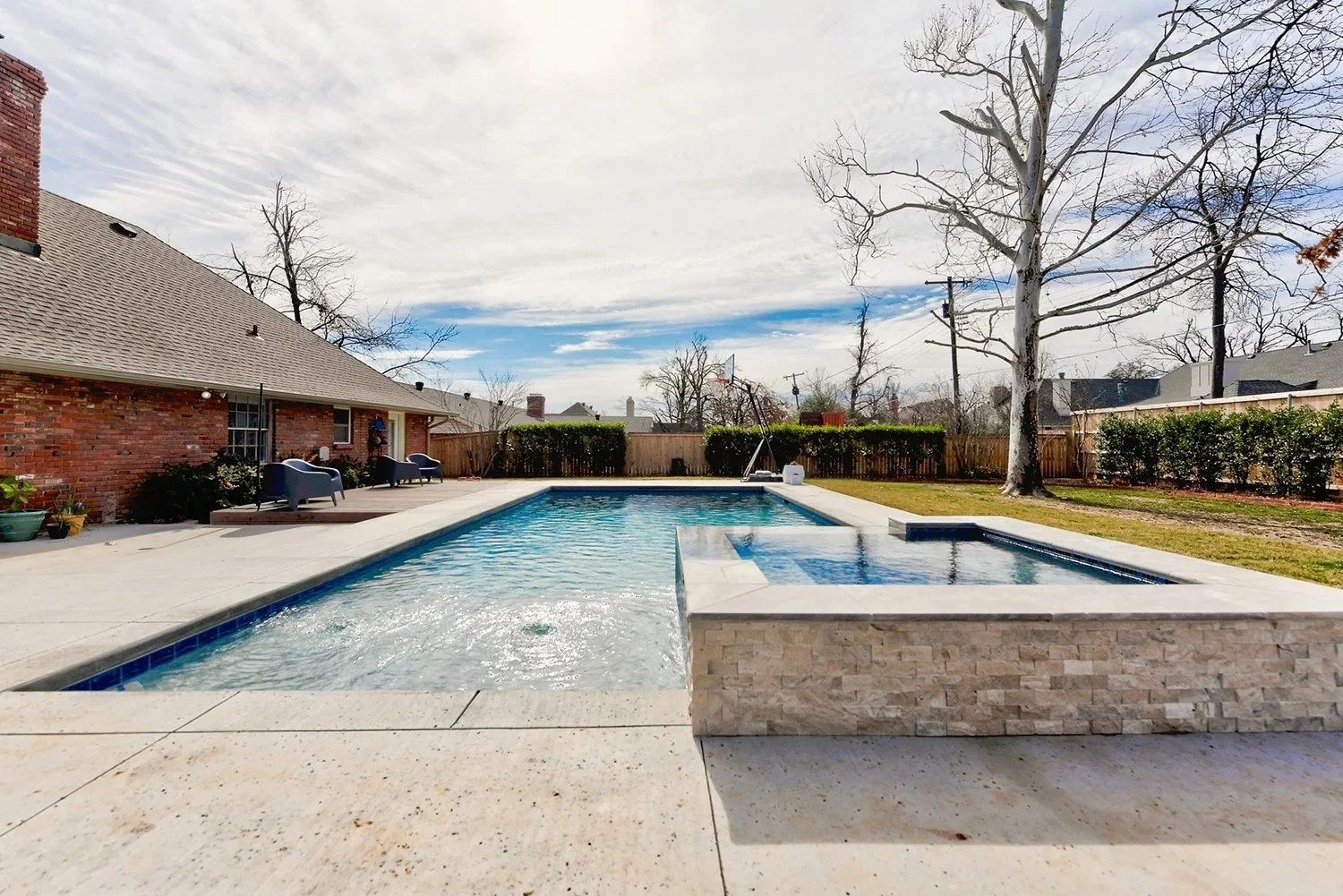 Backyard with swimming pool, hot tub, brick house, leafless trees, and wooden fence under cloudy sky.