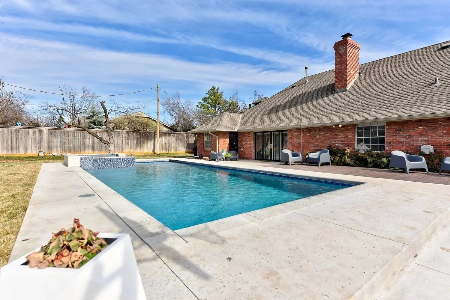 Backyard with swimming pool, concrete patio, red brick house with sliding glass doors, outdoor chairs, and a wooden fence under a blue sky with clouds.