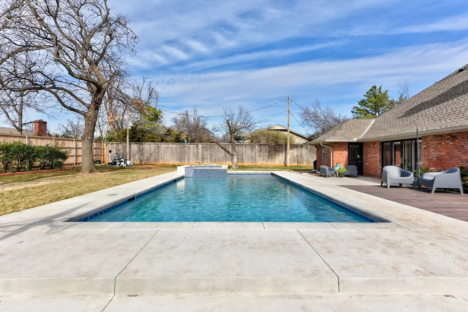 Backyard with a rectangular swimming pool, wooden deck with outdoor chairs, brick house, leafless trees, cloudy blue sky, privacy fence.