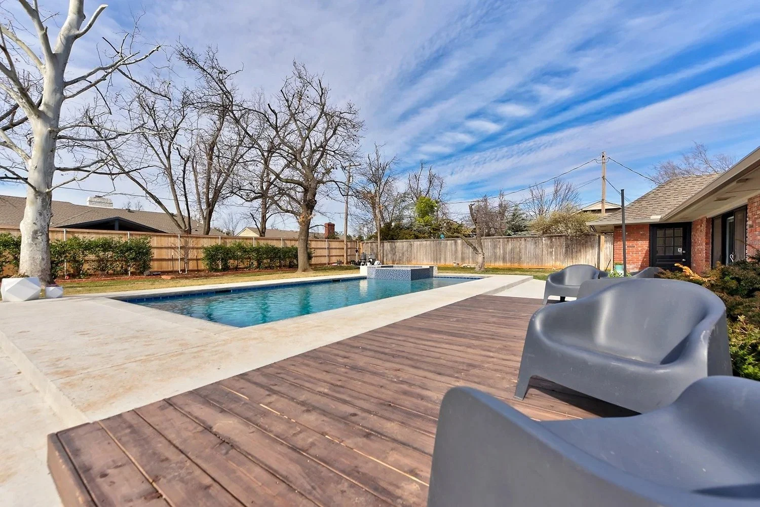 Backyard with swimming pool, pool chairs, and a wooden deck under a blue sky with clouds.