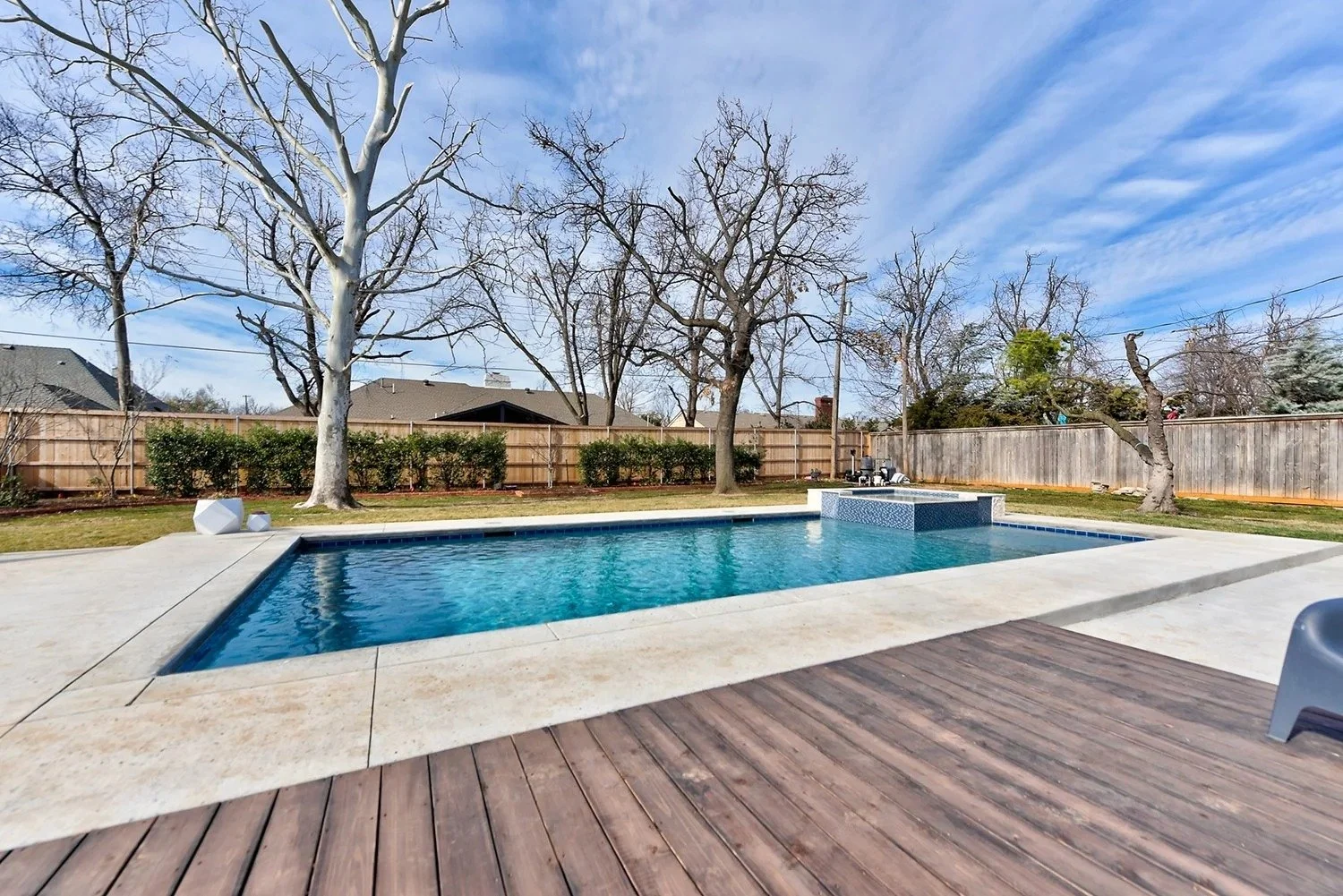 Backyard view of a swimming pool surrounded by a concrete deck and a wooden patio, with trees and a wooden fence in the background, under a partly cloudy sky.