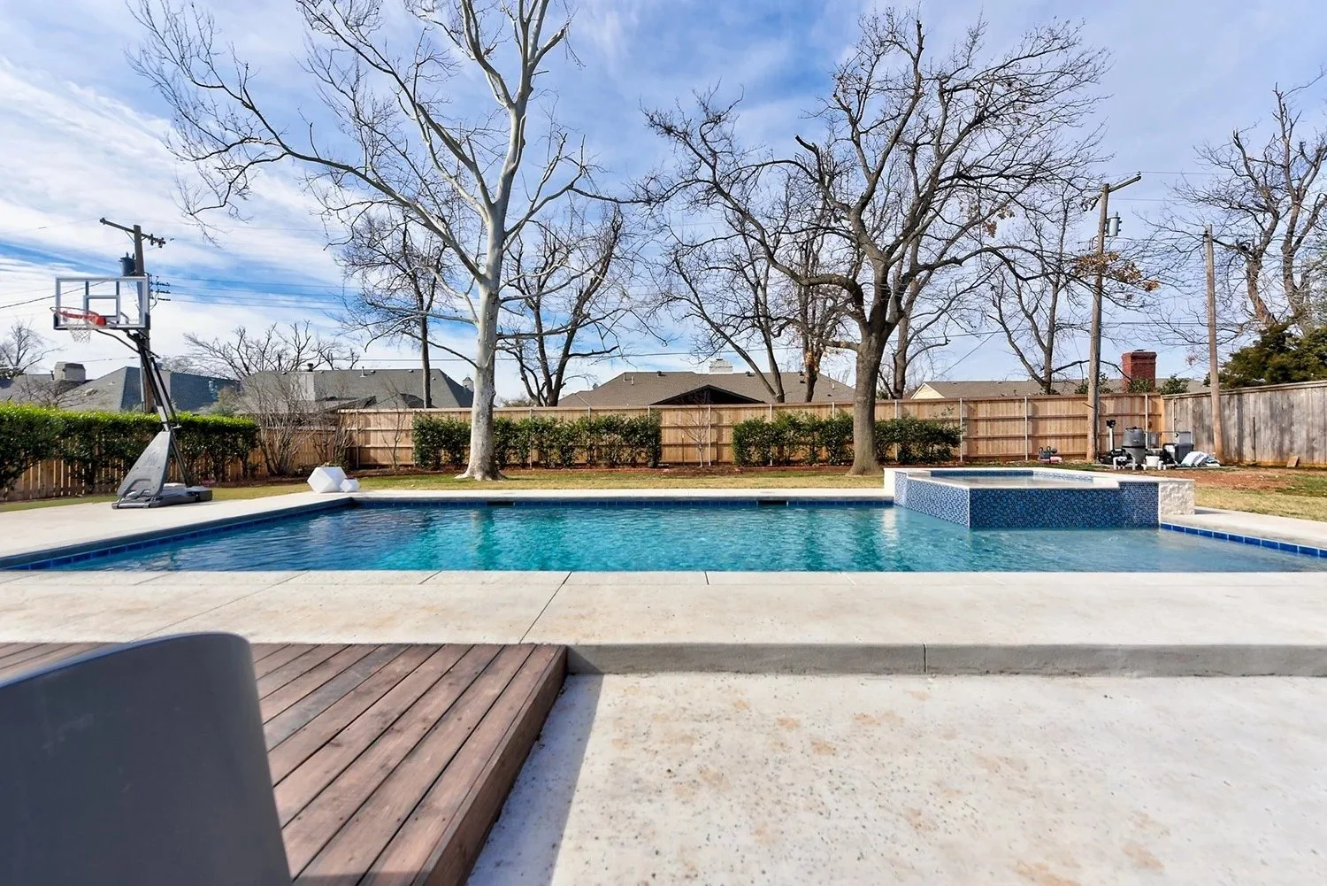 Backyard swimming pool with a basketball hoop and a hot tub, surrounded by a wooden fence and leafless trees, with houses and utility poles in the background on a sunny day
