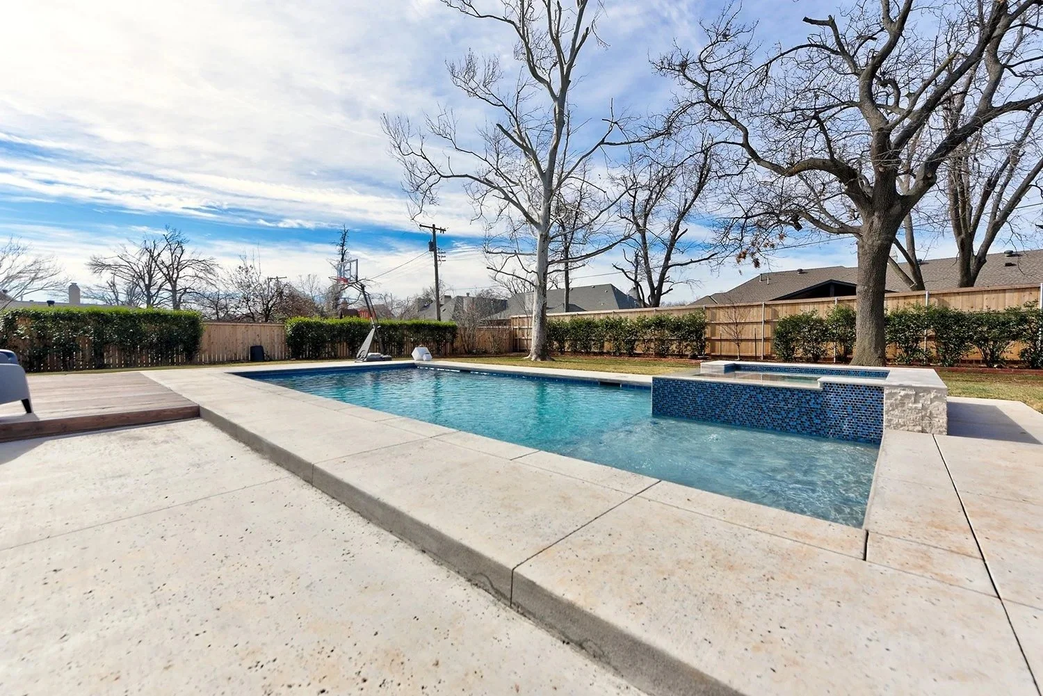 Residential backyard with a rectangular swimming pool, a wooden deck, trees without leaves, a wooden fence, a soccer goal in the distance, and a partly cloudy sky.