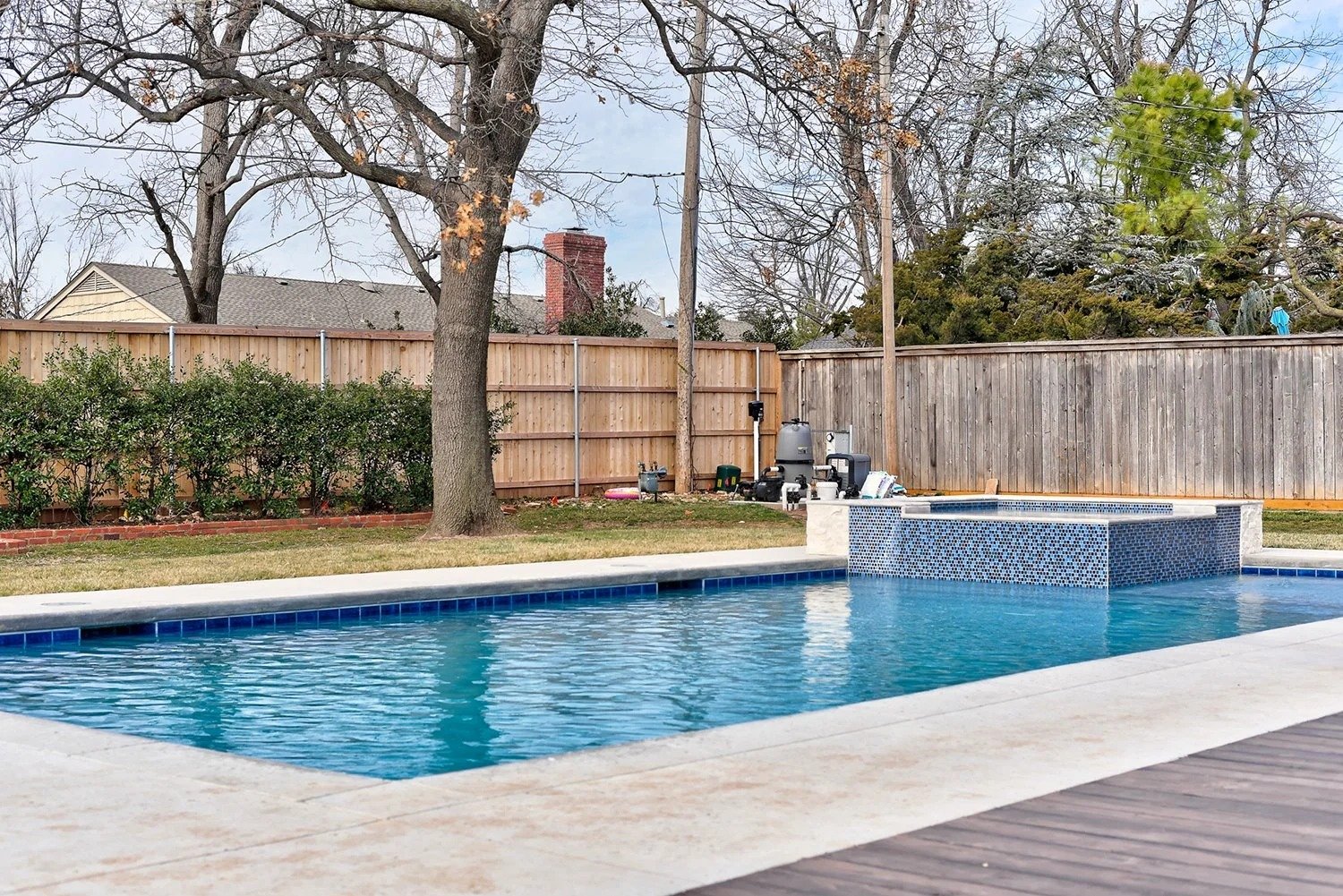 Backyard with a swimming pool, a tall tree, a wooden fence, and some equipment near the fence.