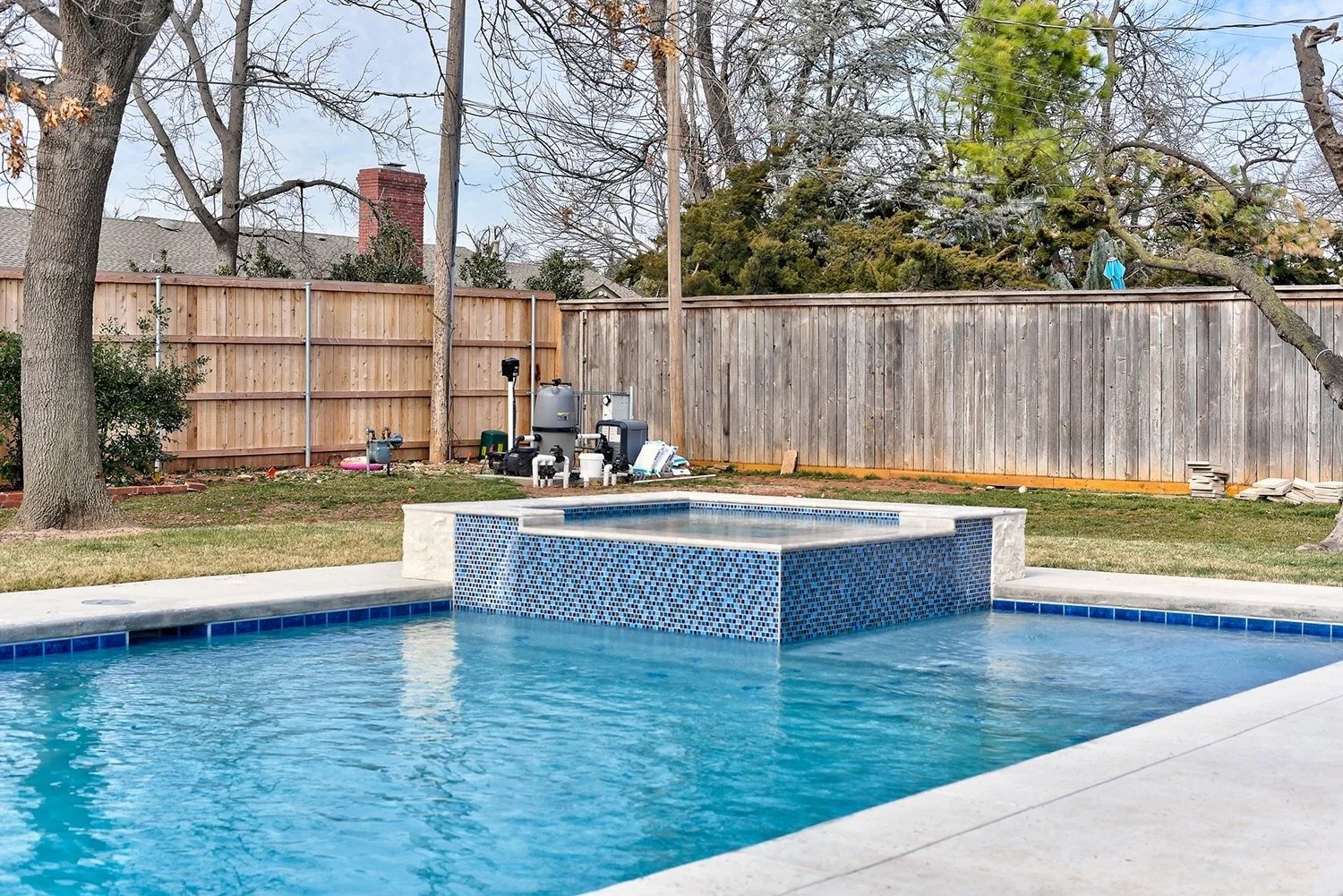 Backyard with a swimming pool featuring a raised hot tub with blue mosaic tiles. Brown and gray wooden fences surround the yard. There are some trees, one with branches extending over the fence. Equipment and supplies are placed near the fence, inclu