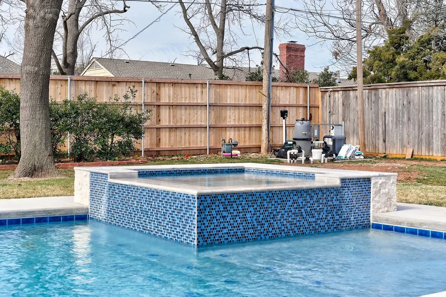 Backyard with a swimming pool and a raised hot tub with blue mosaic tiles, surrounded by a concrete deck, tall wooden fence, a large tree, and various pool equipment in the background.