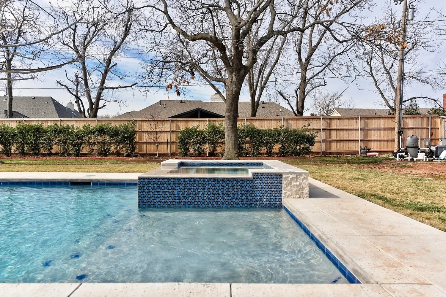 Backyard with a swimming pool, a tree in the center, a wooden fence, neighboring houses, and some outdoor equipment on the right.