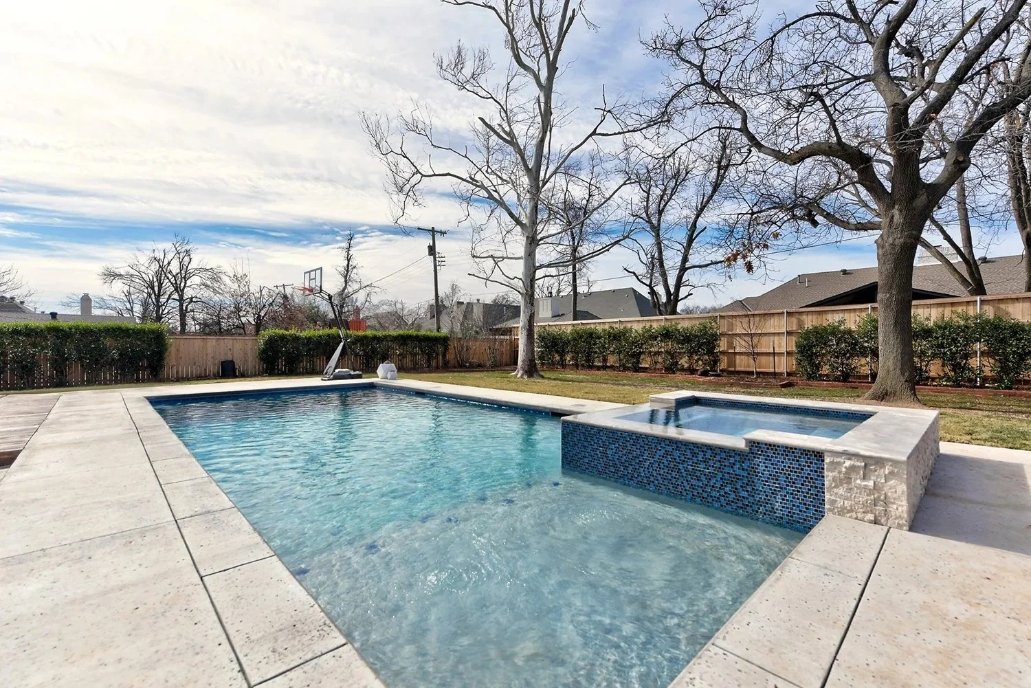 Backyard with swimming pool, hot tub, fenced yard, trees, and partly cloudy sky.