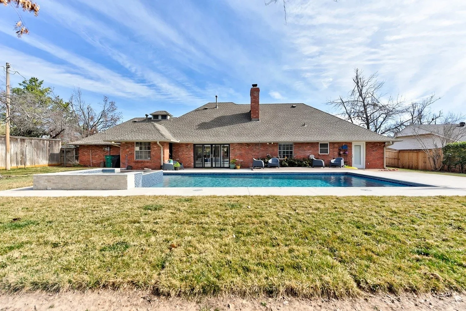Backyard of a brick house with a swimming pool, patio furniture, and a grassy lawn, surrounded by a wooden fence and trees.