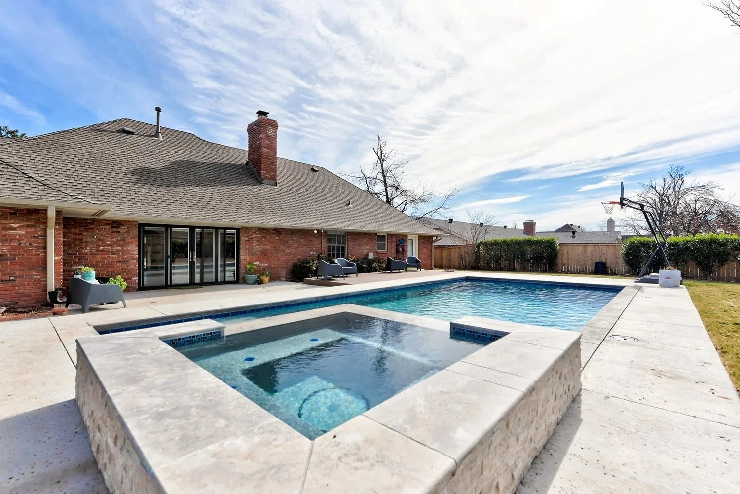 Backyard with swimming pool, hot tub, patio chairs, and a basketball hoop, fenced yard, brick house with sliding glass doors, clear sky with some clouds.