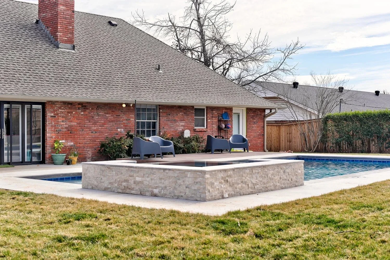 Backyard patio area with a swimming pool, two chairs, a fire pit, potted plants, and a brick house with a glass sliding door.
