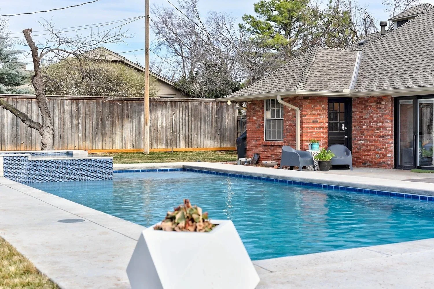 Residential backyard featuring a swimming pool with blue mosaic tile accents, a concrete patio, a brick house with a shingled roof, outdoor furniture, potted plants, a wooden fence, trees, and overcast sky.