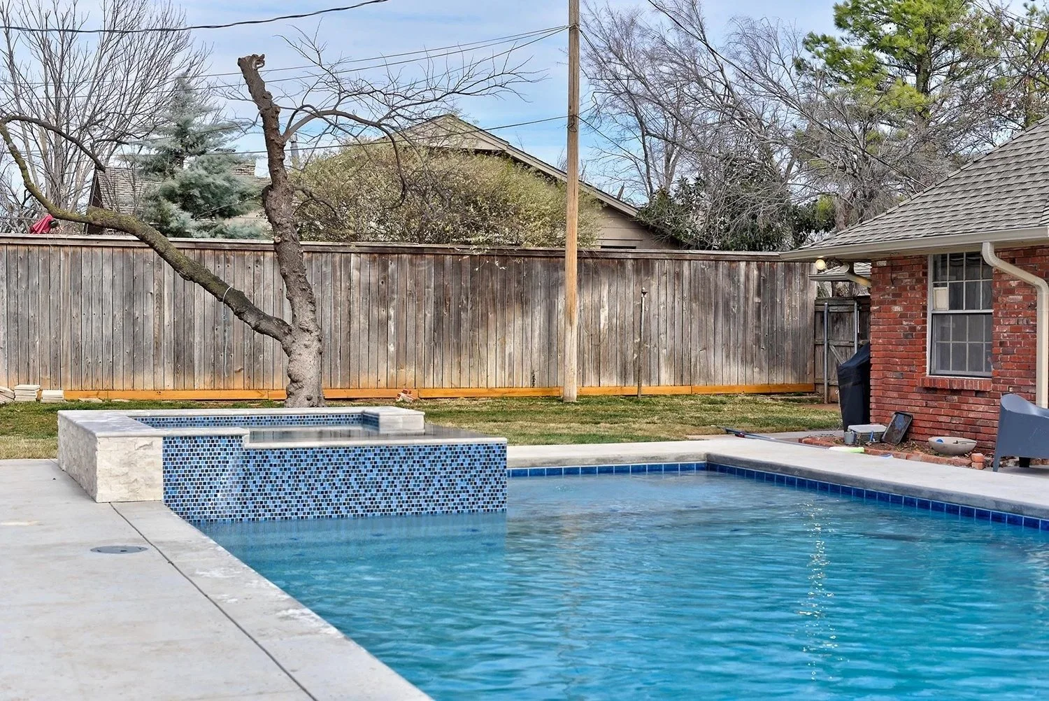 Residential backyard with a swimming pool and hot tub, wooden fence, trees, and a brick house with a window.