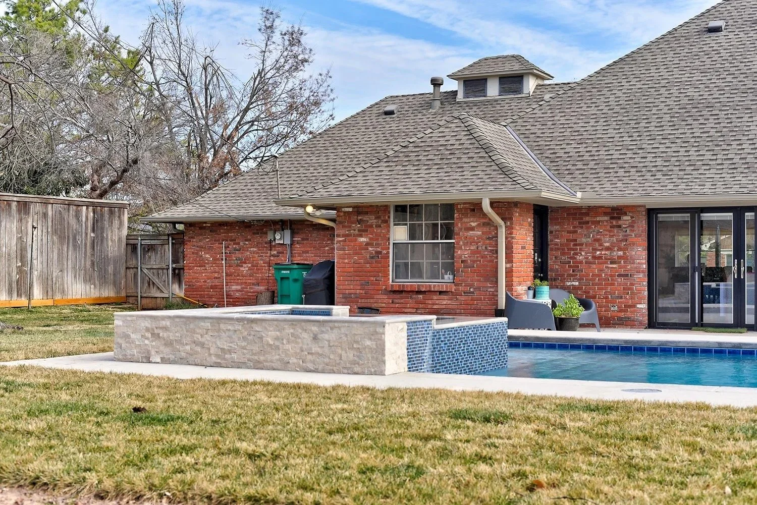 Backyard with a brick house, swimming pool, and a stone hot tub surrounded by a grassy yard, wooden fence, and trees in the background.