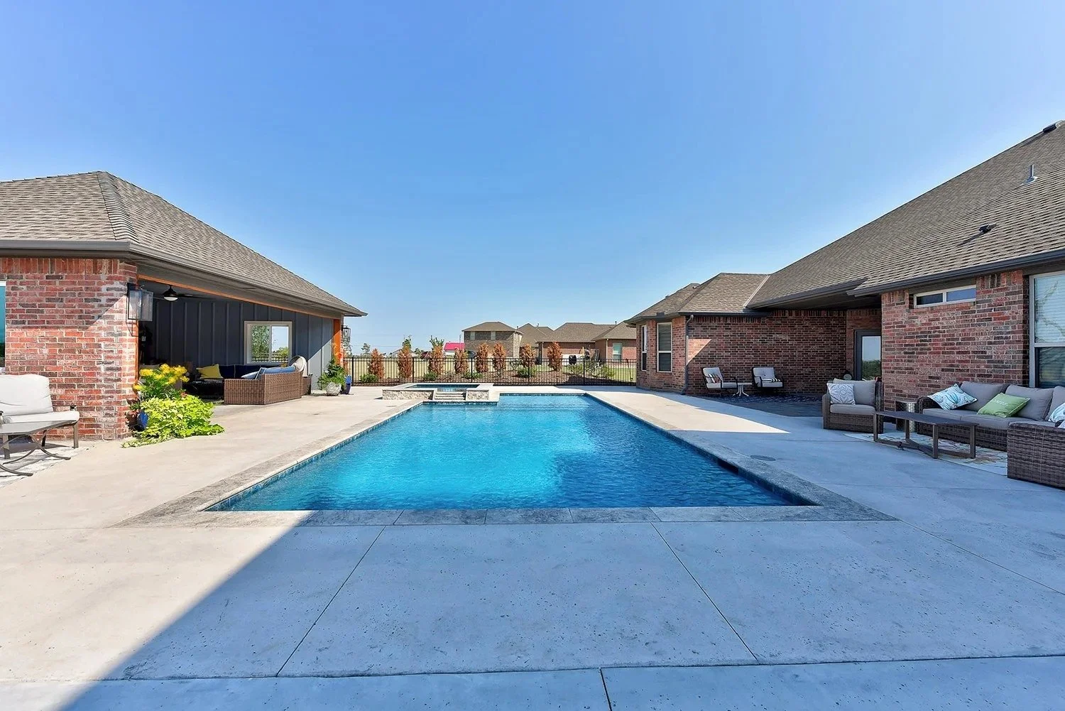 A backyard with a swimming pool surrounded by concrete patio, with outdoor seating and a fence with trees in the background.