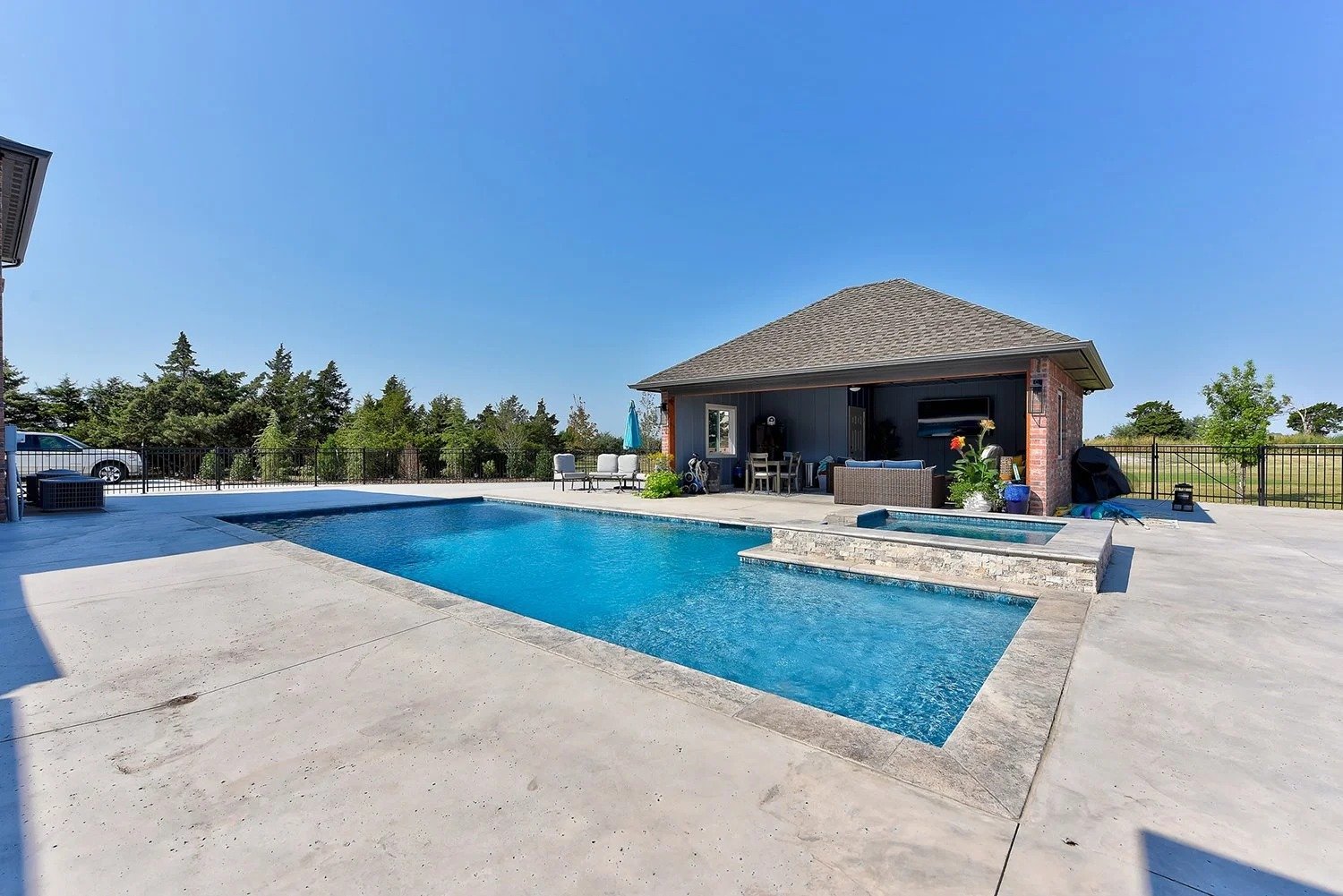 A backyard swimming pool with clear blue water, facing a pool house with outdoor furniture, potted plants, and a barbecue grill on a sunny day with a bright blue sky.